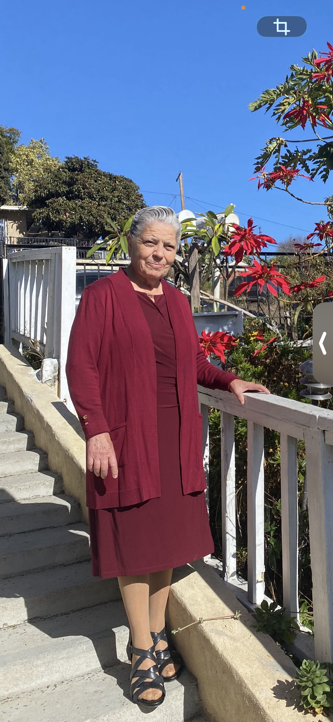Woman stands on a walkway beside a garden filled with colorful flowers in a residential setting.