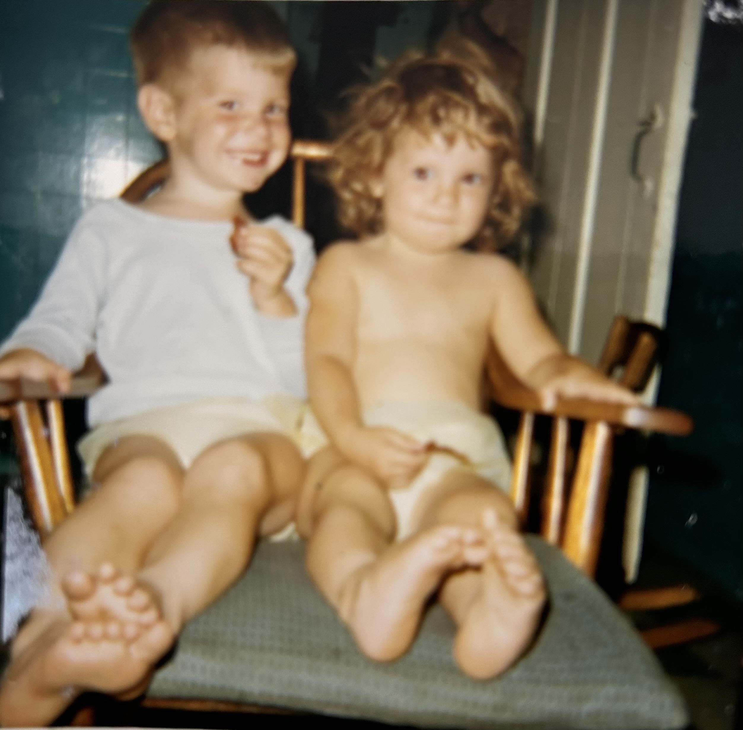 Two young children with light skin sit on a chair in a home. They look at the camera.
