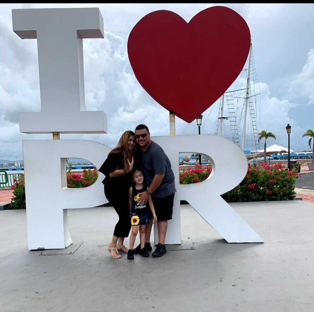 Family poses together at a large love sign in Puerto Rico by the waterfront with trees and boats.