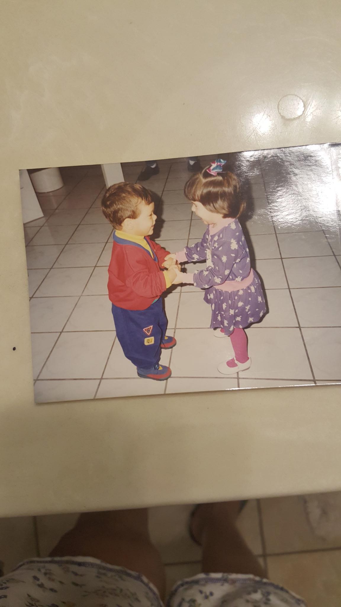 Two kids hold hands and smile while playing in a classroom with tiled floors.