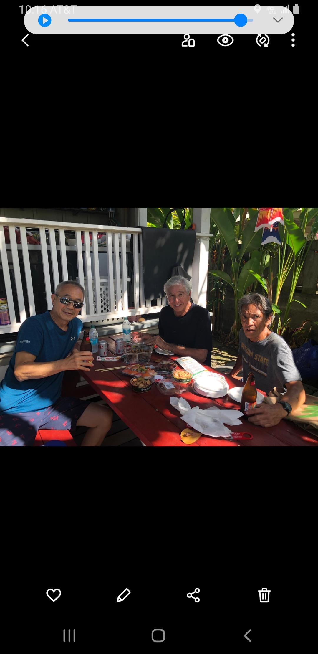 Three friends gather around a red table filled with food and drinks on a sunny day.