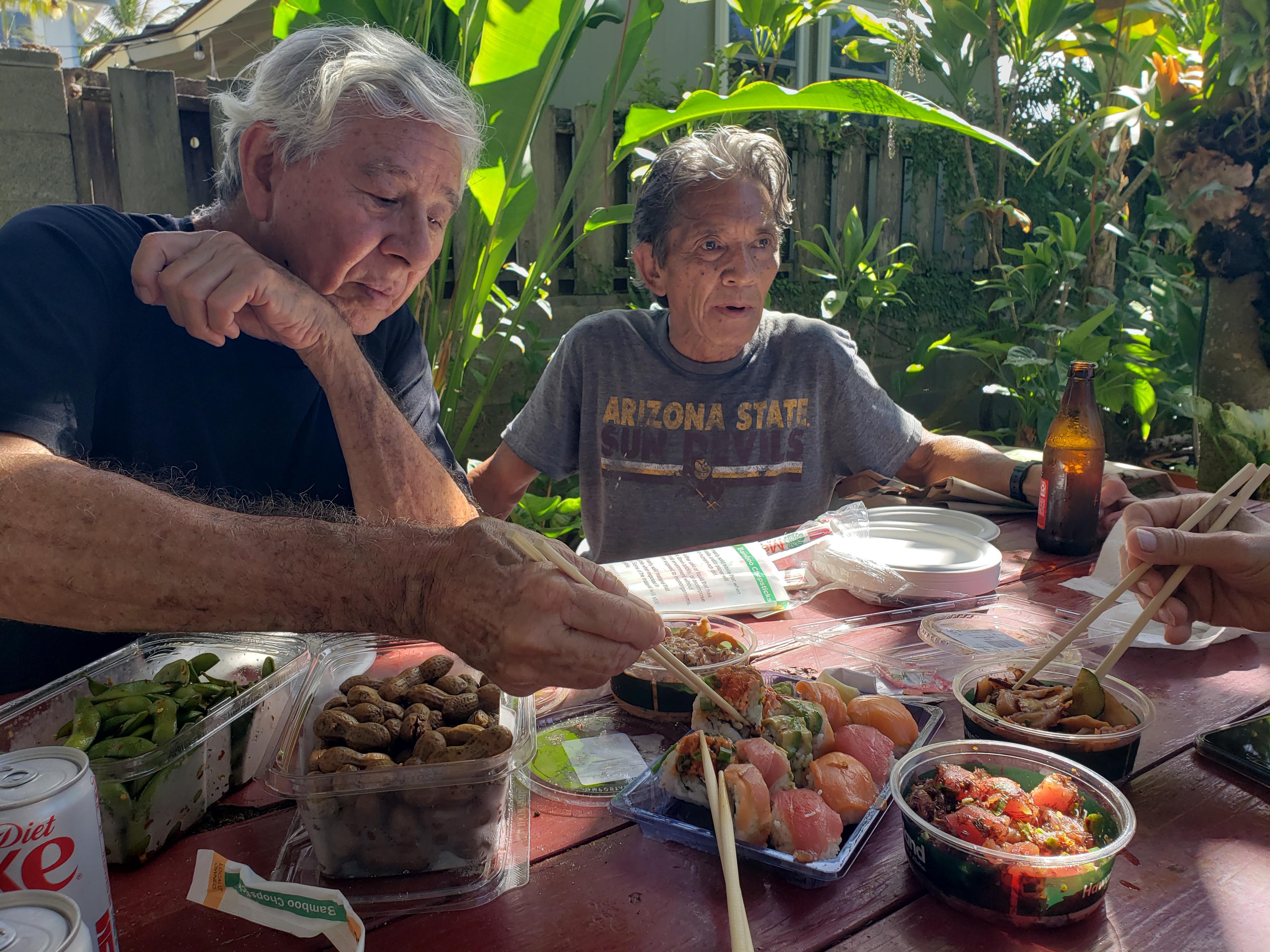 Two men share a meal together at a table surrounded by plants and greenery during the day.