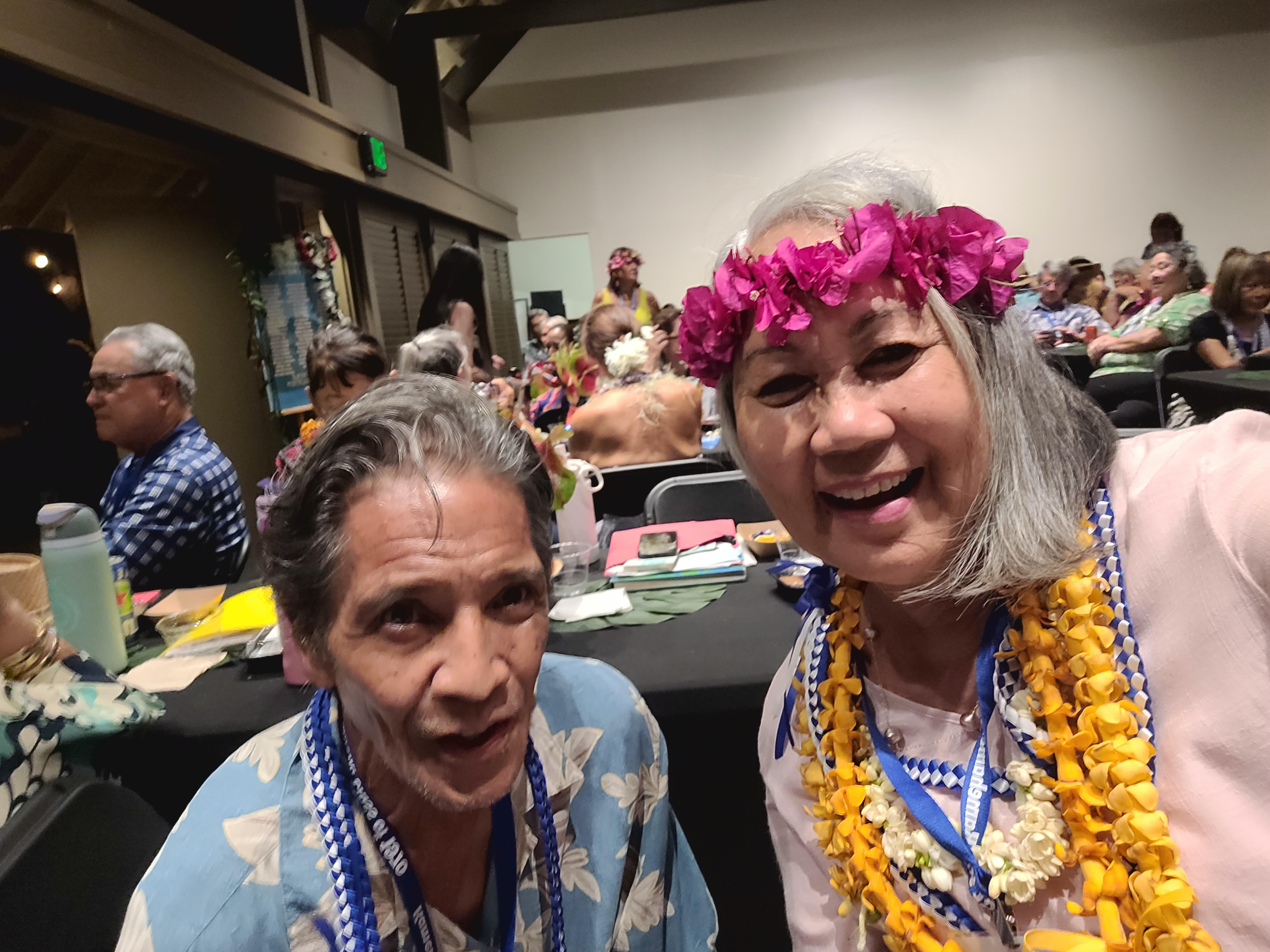 Two individuals smile at the camera while surrounded by others at a cultural gathering indoors.