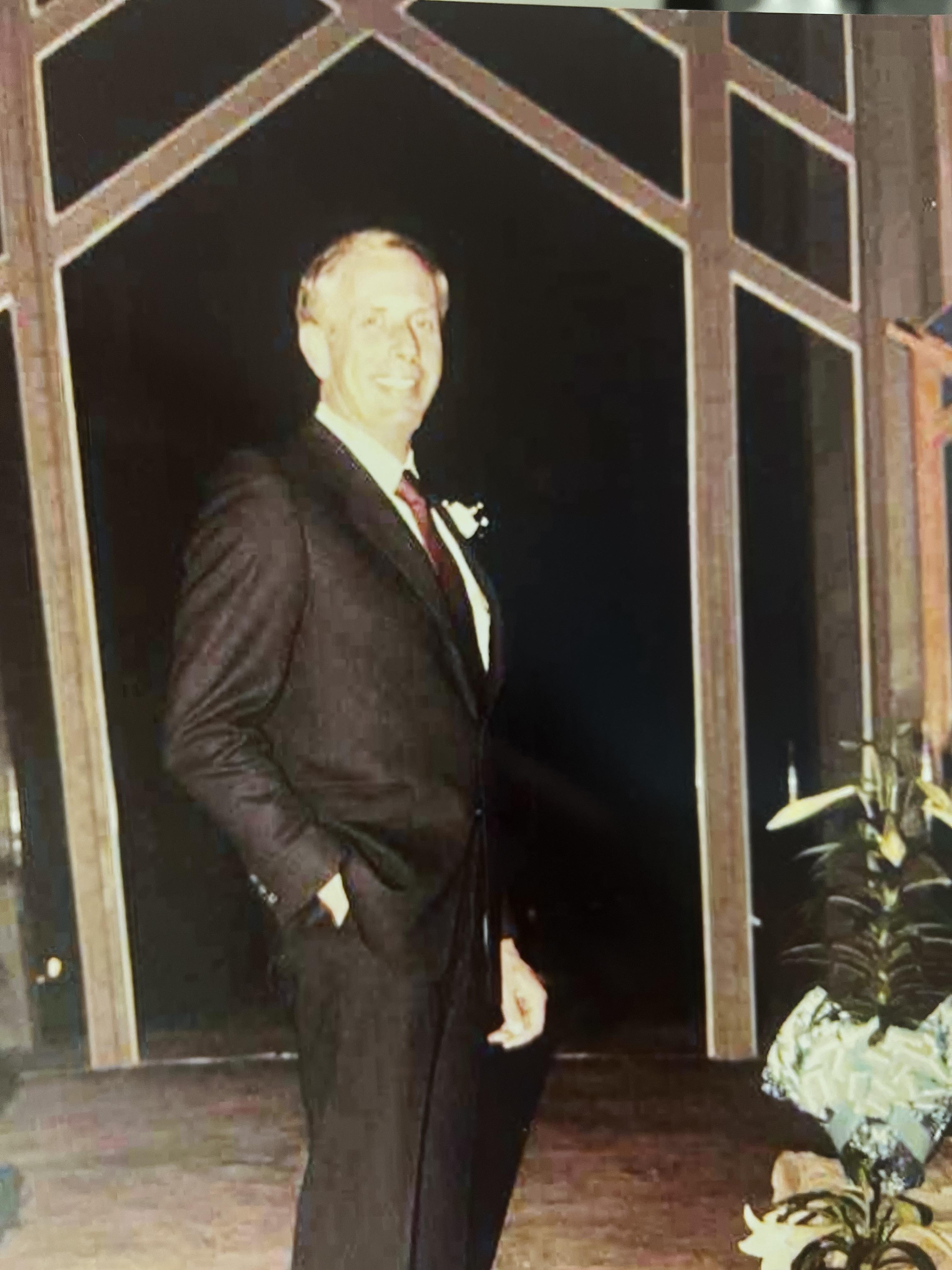 A man poses near a window wearing a suit at a social gathering during evening hours.