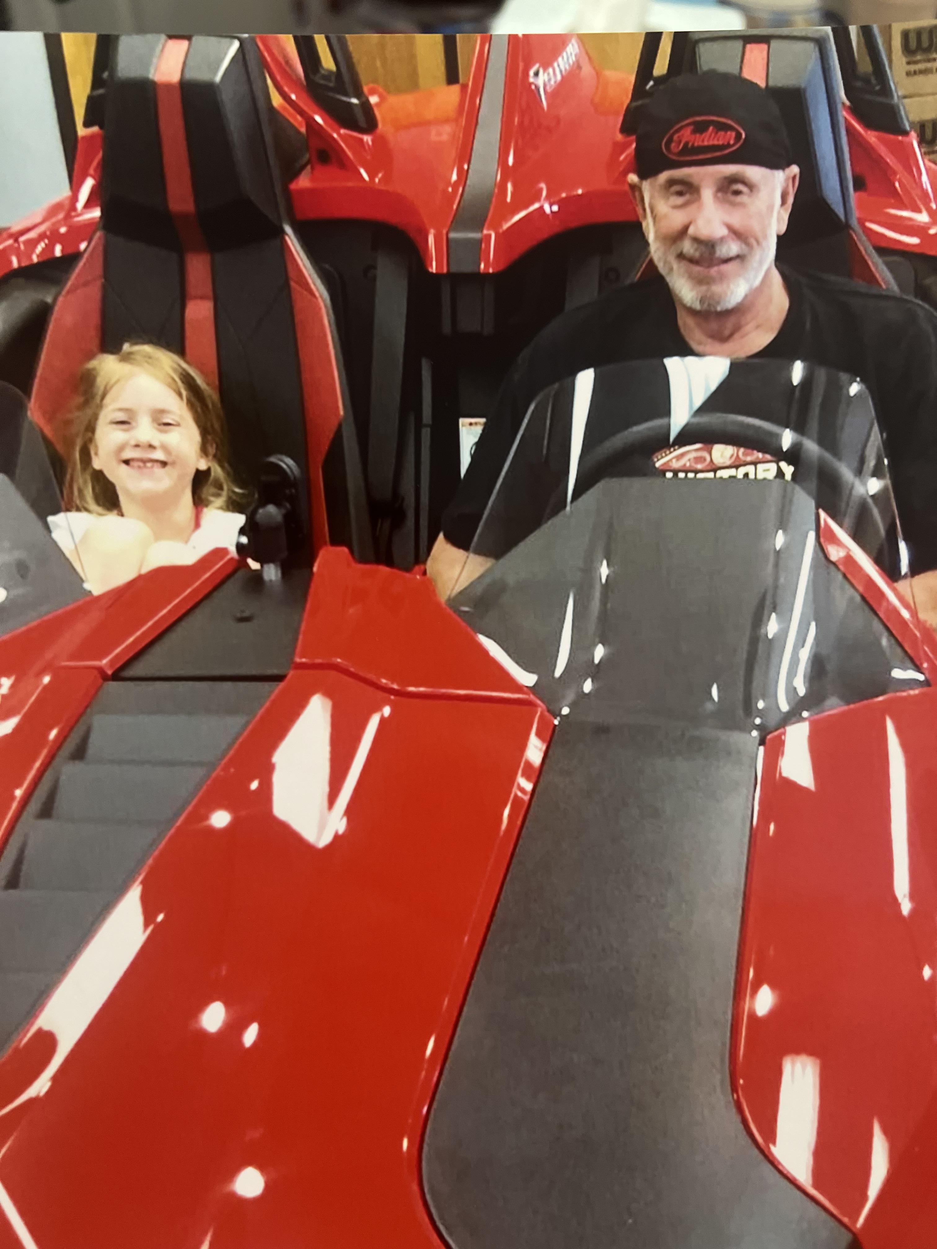 A man and a girl smile while sitting in a red race car at an indoor event in the city.