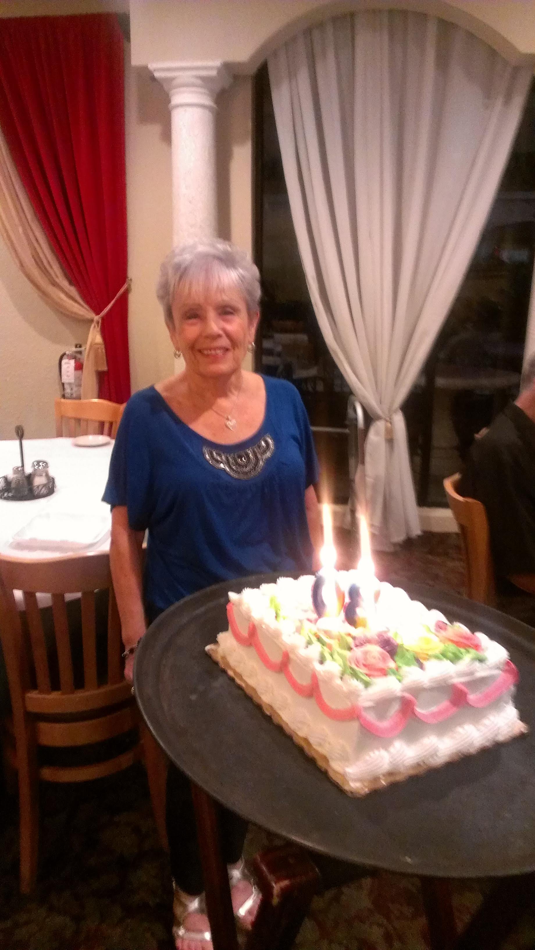 A woman stands beside a birthday cake with lit candles in a restaurant setting.