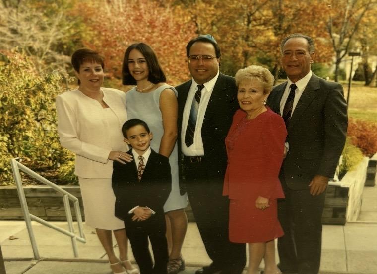 Family stands together in a park, trees show autumn colors. Everyone smiles at the camera.