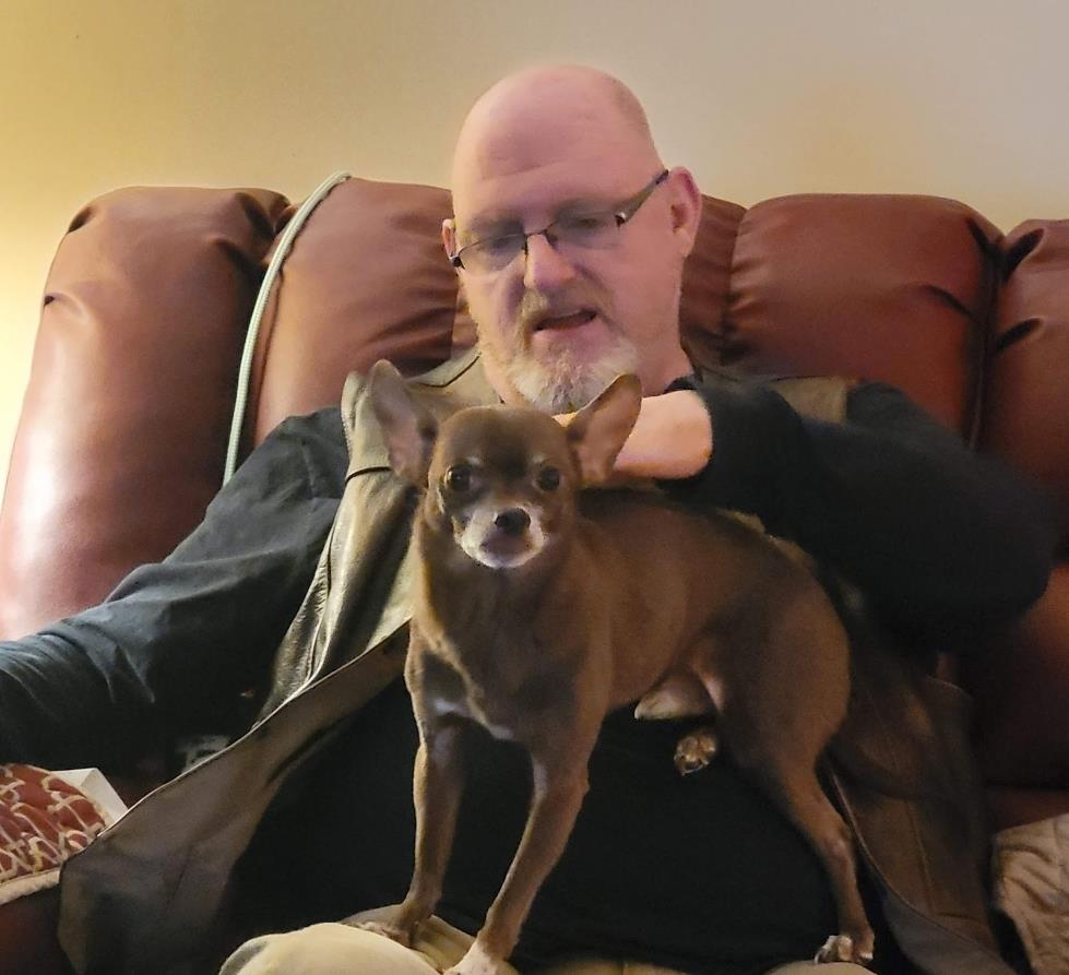 A man sits on a couch while holding a small dog on his lap at home in the afternoon.