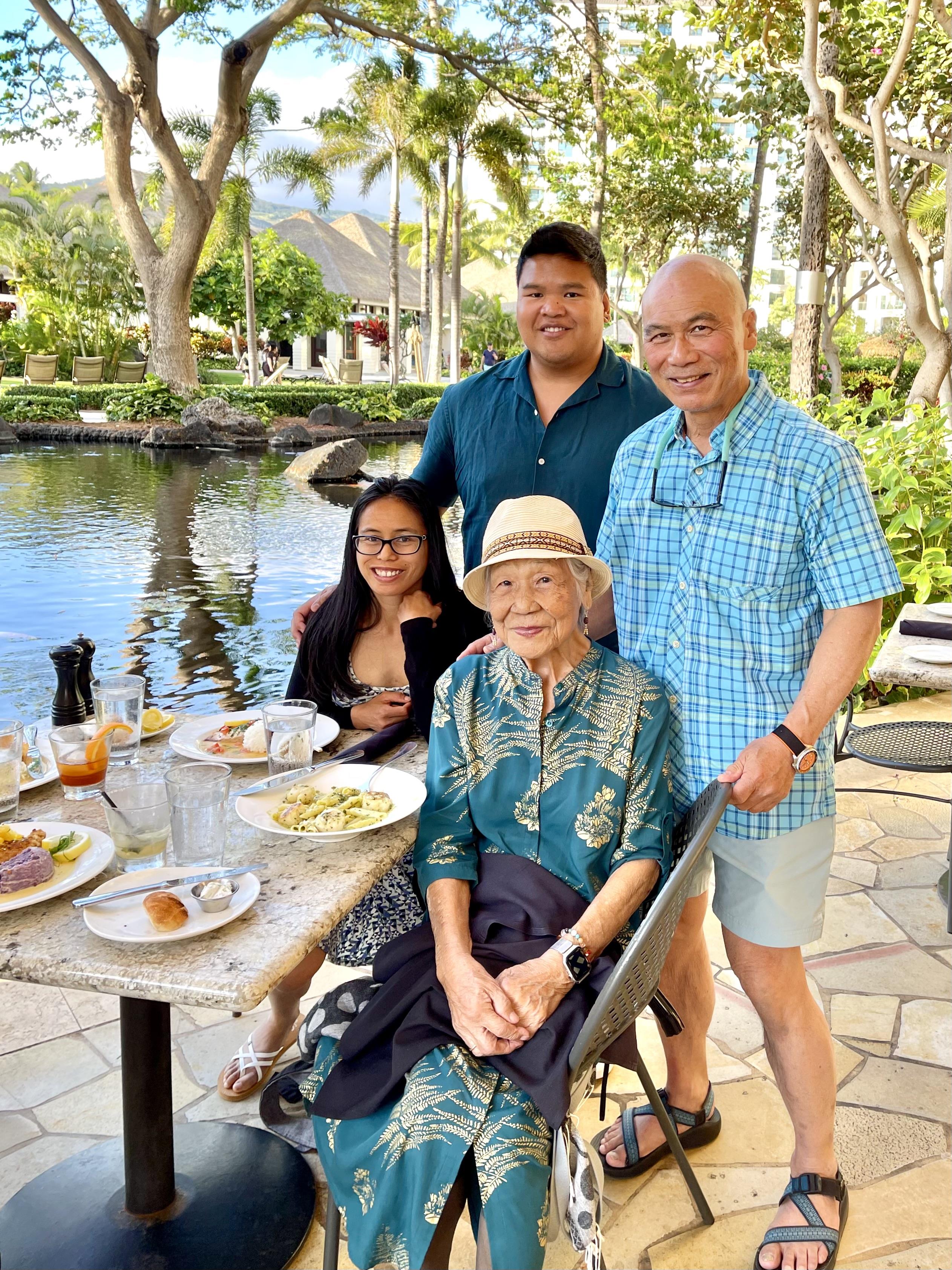 Family sits around a table near the water, sharing food and smiling together in the sun.
