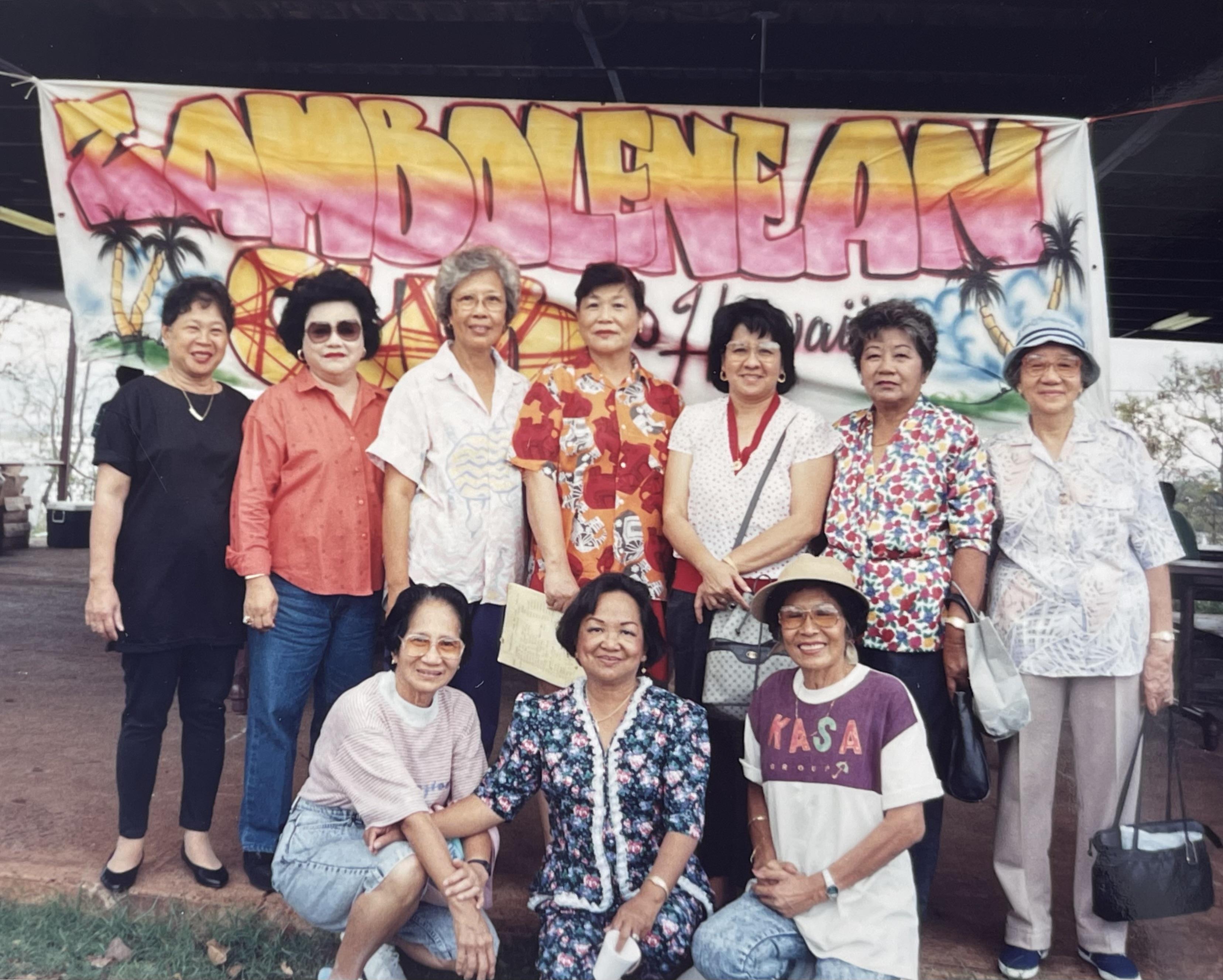 A group of women stands together in Hawaii at a cultural event, smiling at the camera.