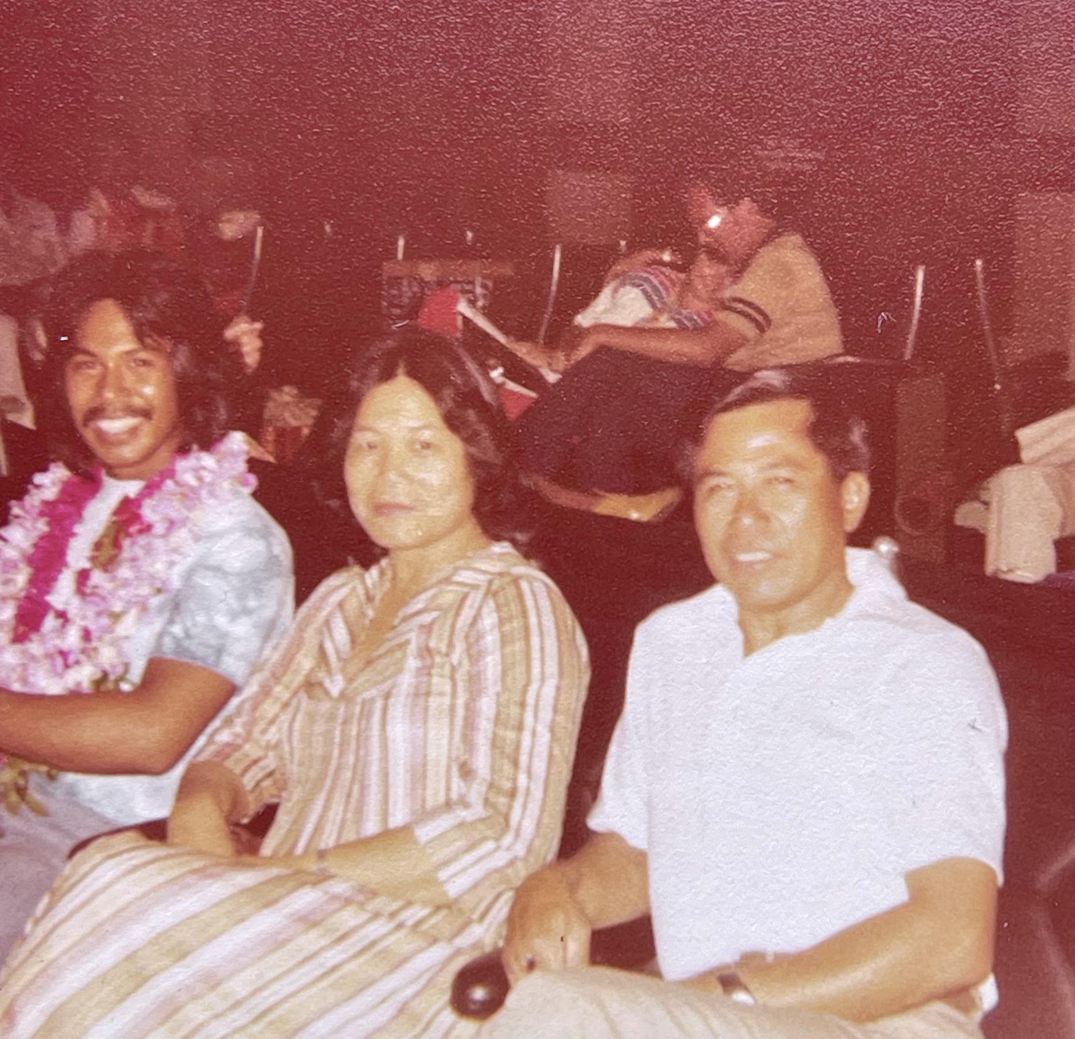 Three individuals sit together in a hall during a social event in the 1970s.