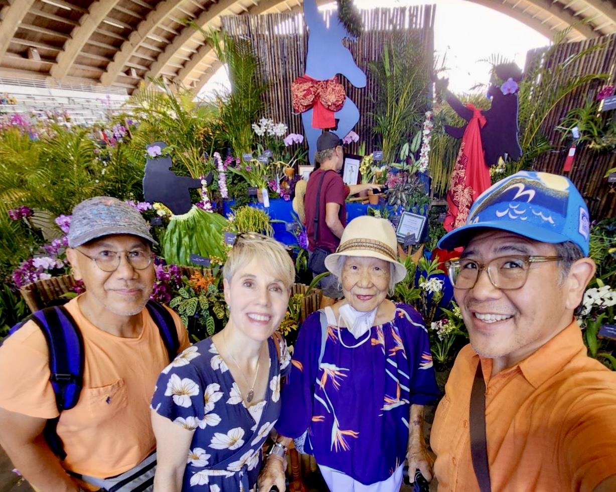 Four people stand close together, smiling at a flower display in a market with colorful plants.