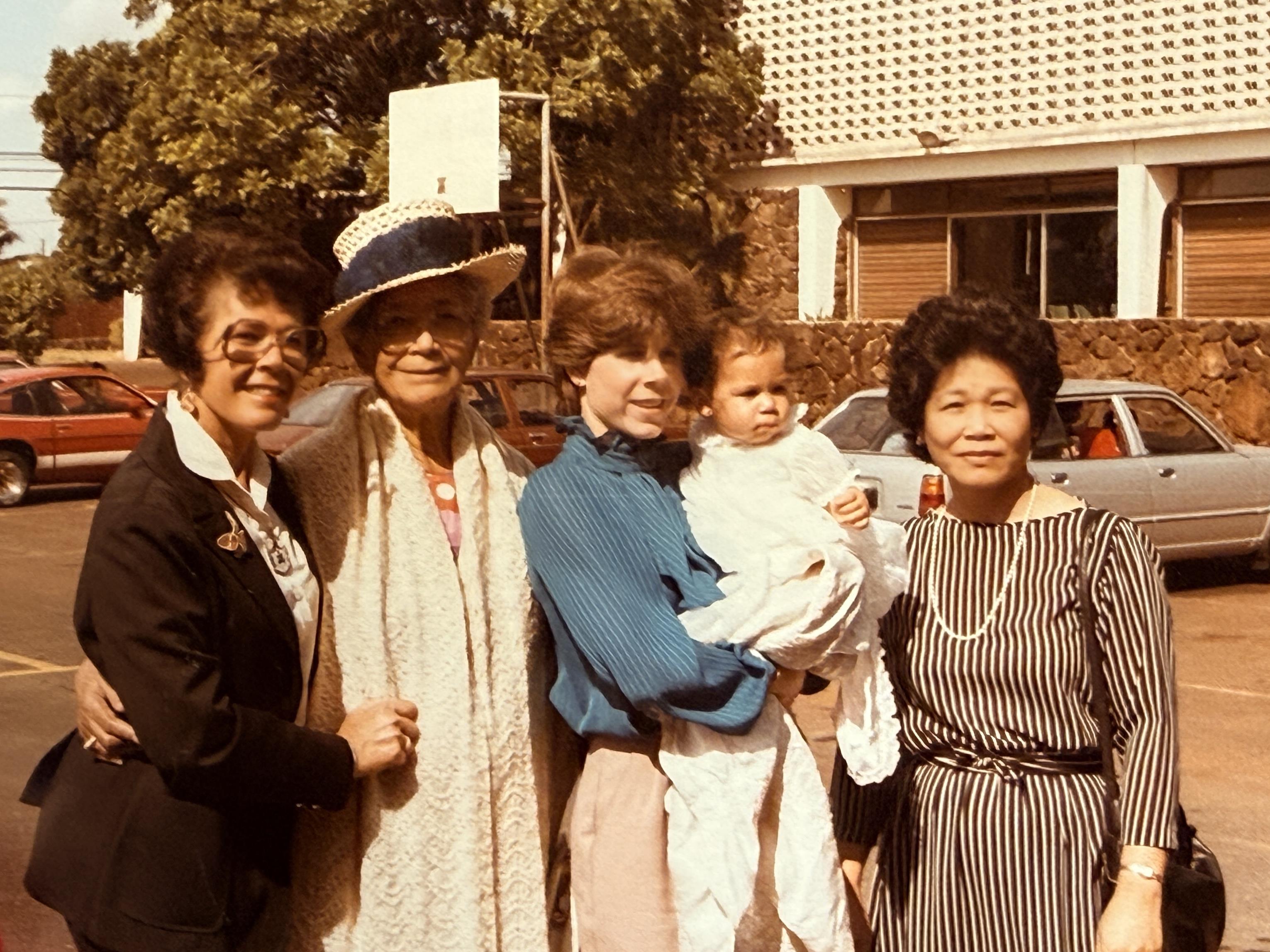 Five women stand together outside near cars with one holding a baby. The sun is shining bright.