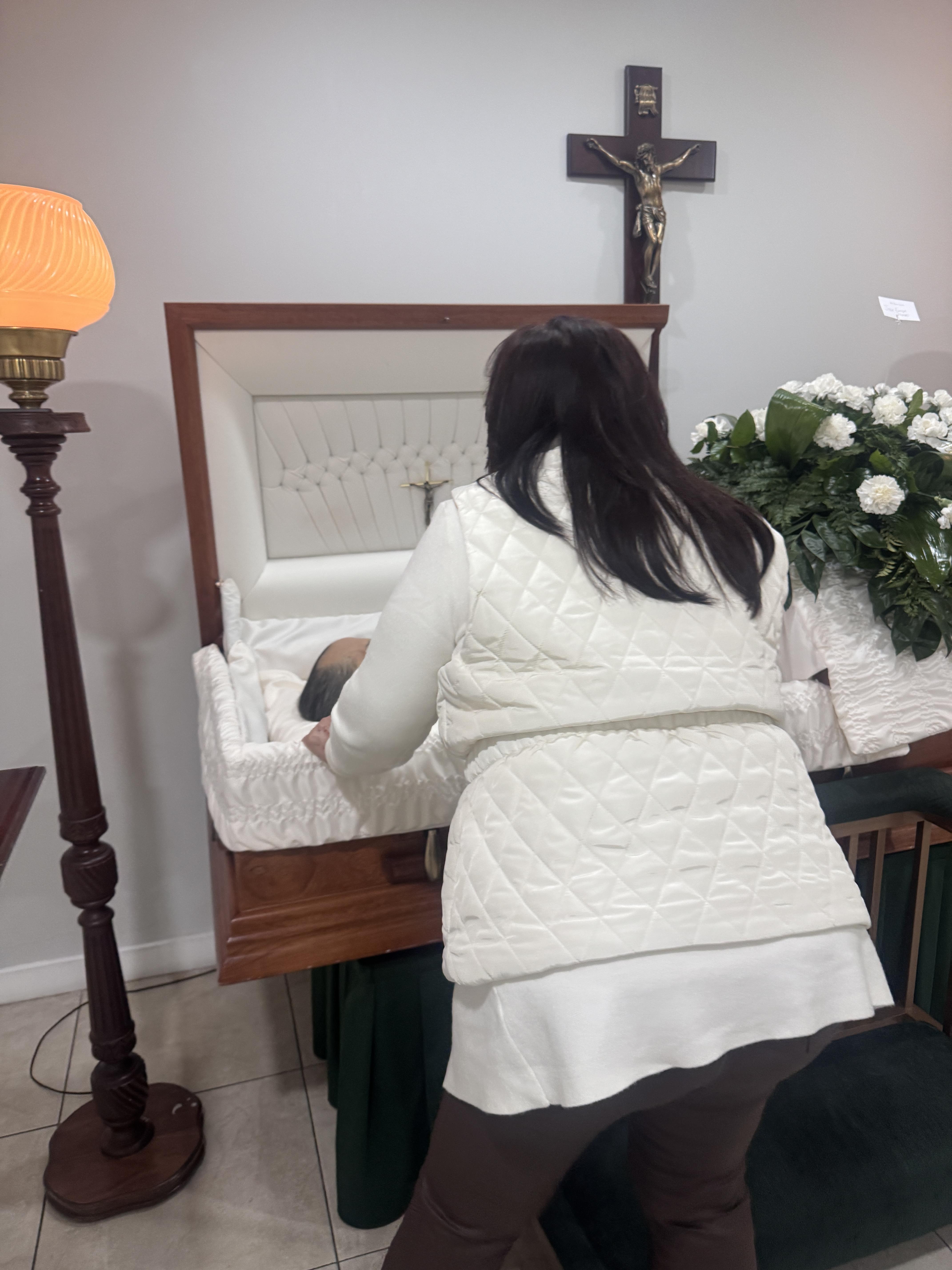 A person bows over a casket during a funeral service at a small room filled with flowers.