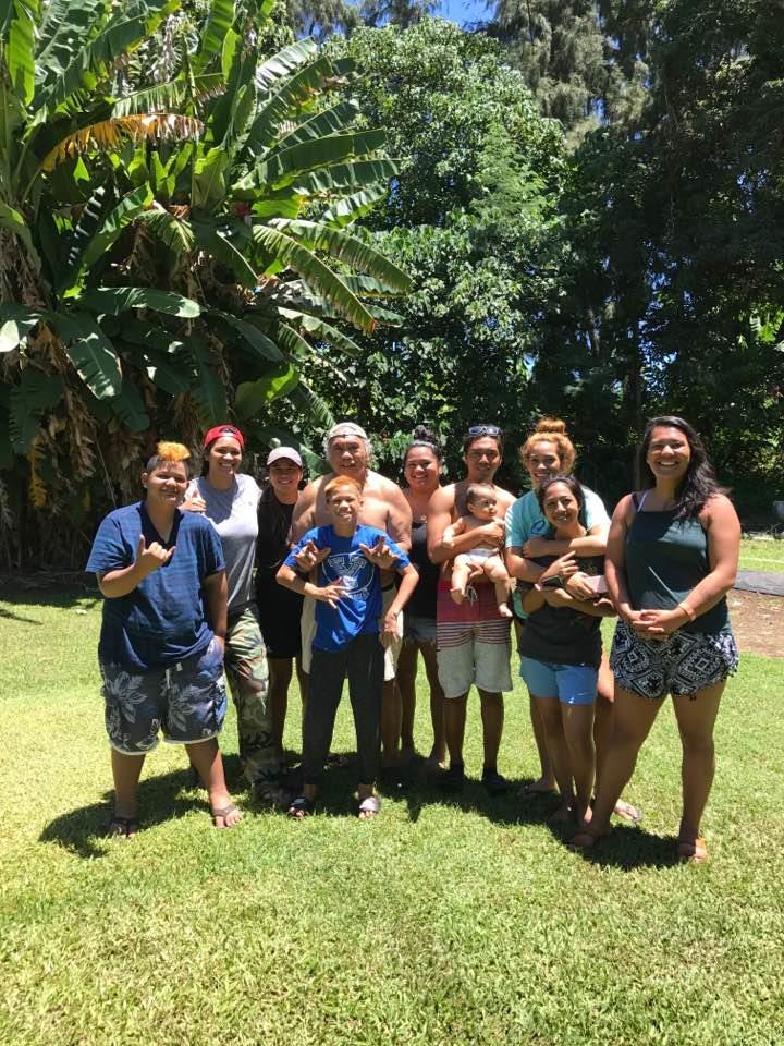 A group of ten people stands together on grass in a sunny location by trees, smiling and posing.