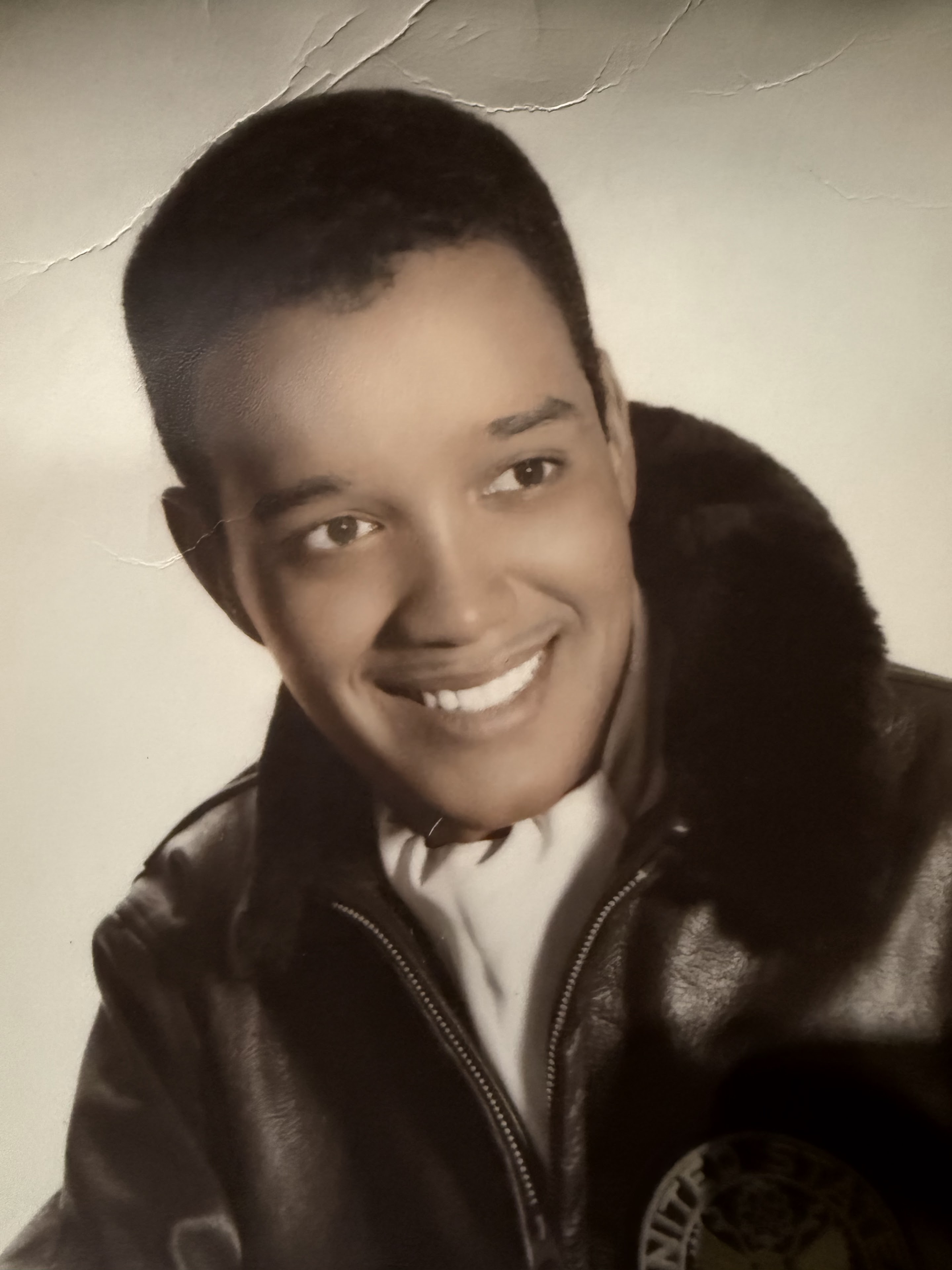 A man is smiling while wearing a leather jacket with fur trim on a light background.