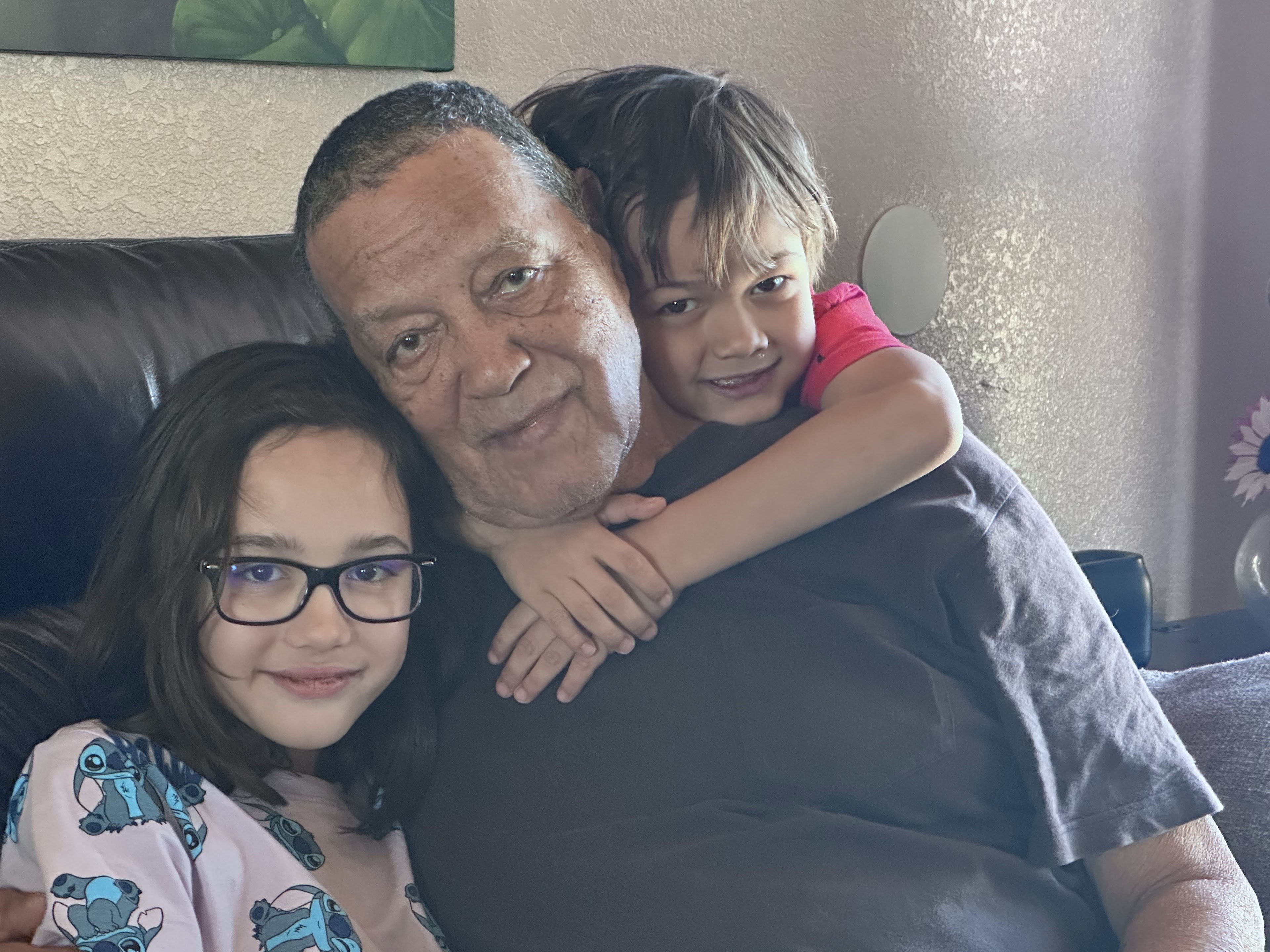 Two children hug an older man while smiling in a cozy living room during the day.