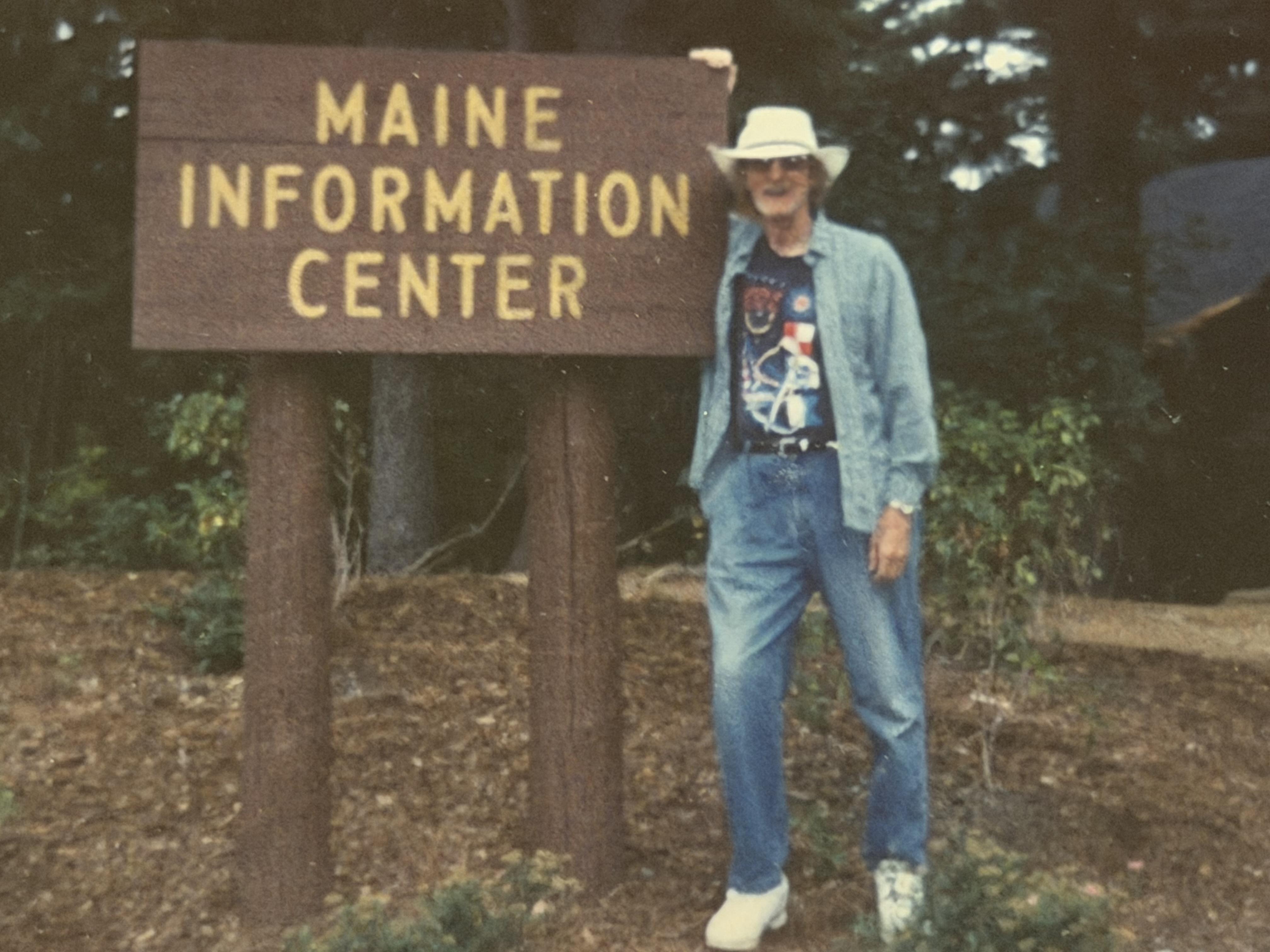 A man poses by the Maine Information Center sign while wearing casual clothes and a hat.