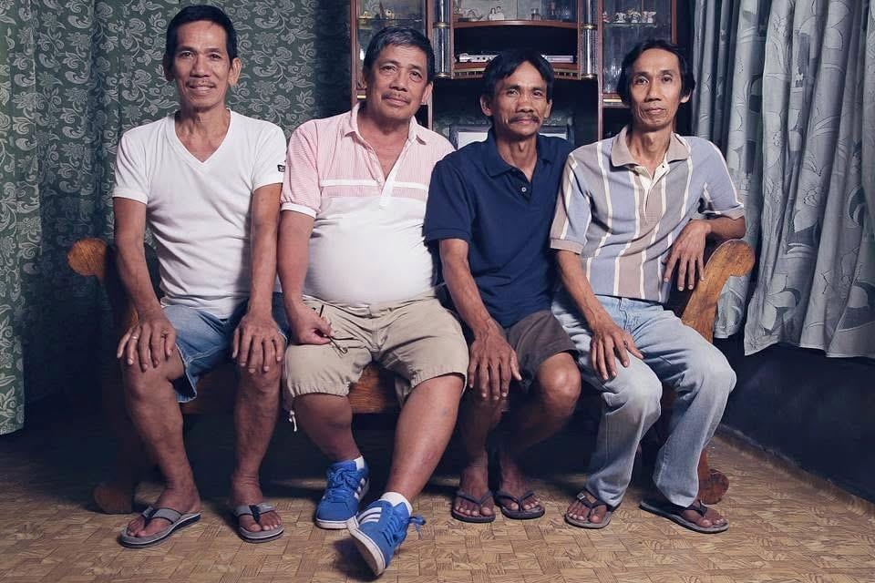 Four men sit on a bench with relaxed expressions, enjoying each other's company while indoors.