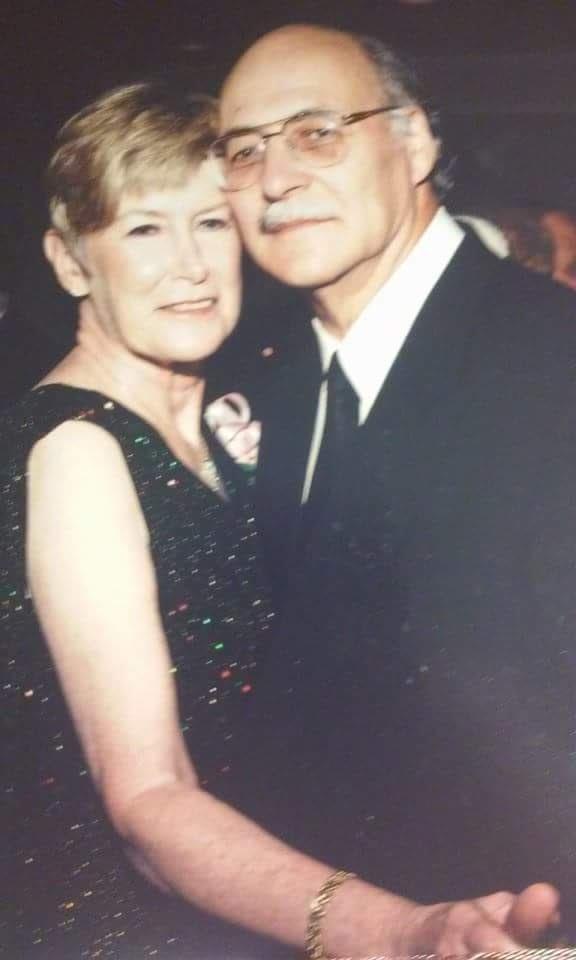 A couple stands close together in formal attire at a social gathering in the evening.