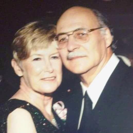 A couple stands closely together, smiling, at a social gathering during the evening.