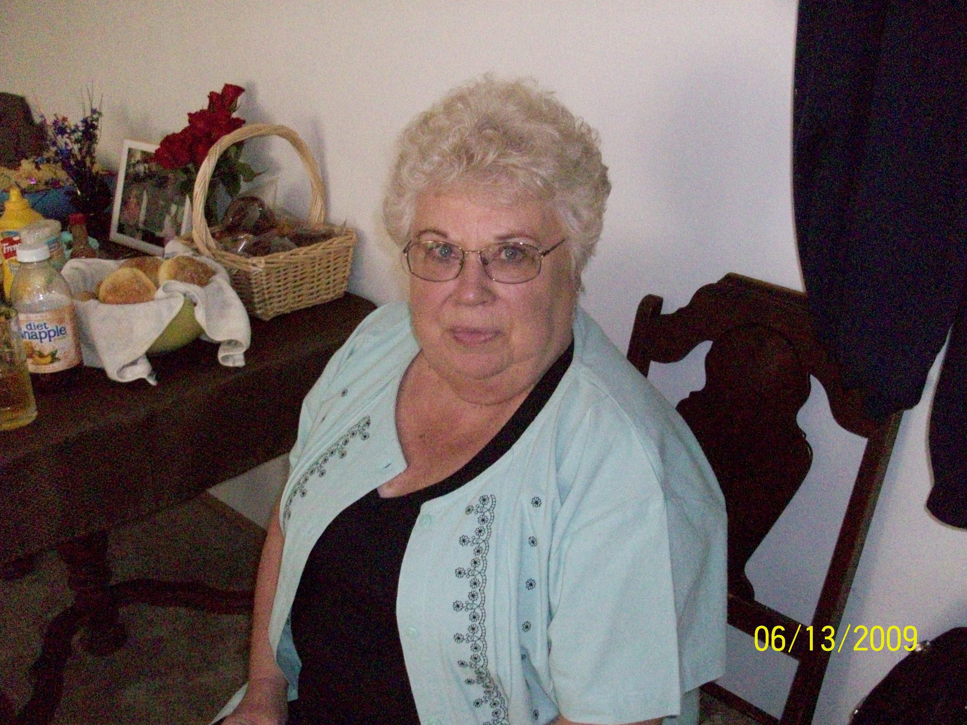 A woman with glasses sits at a table with a basket and other items nearby during a social gathering.