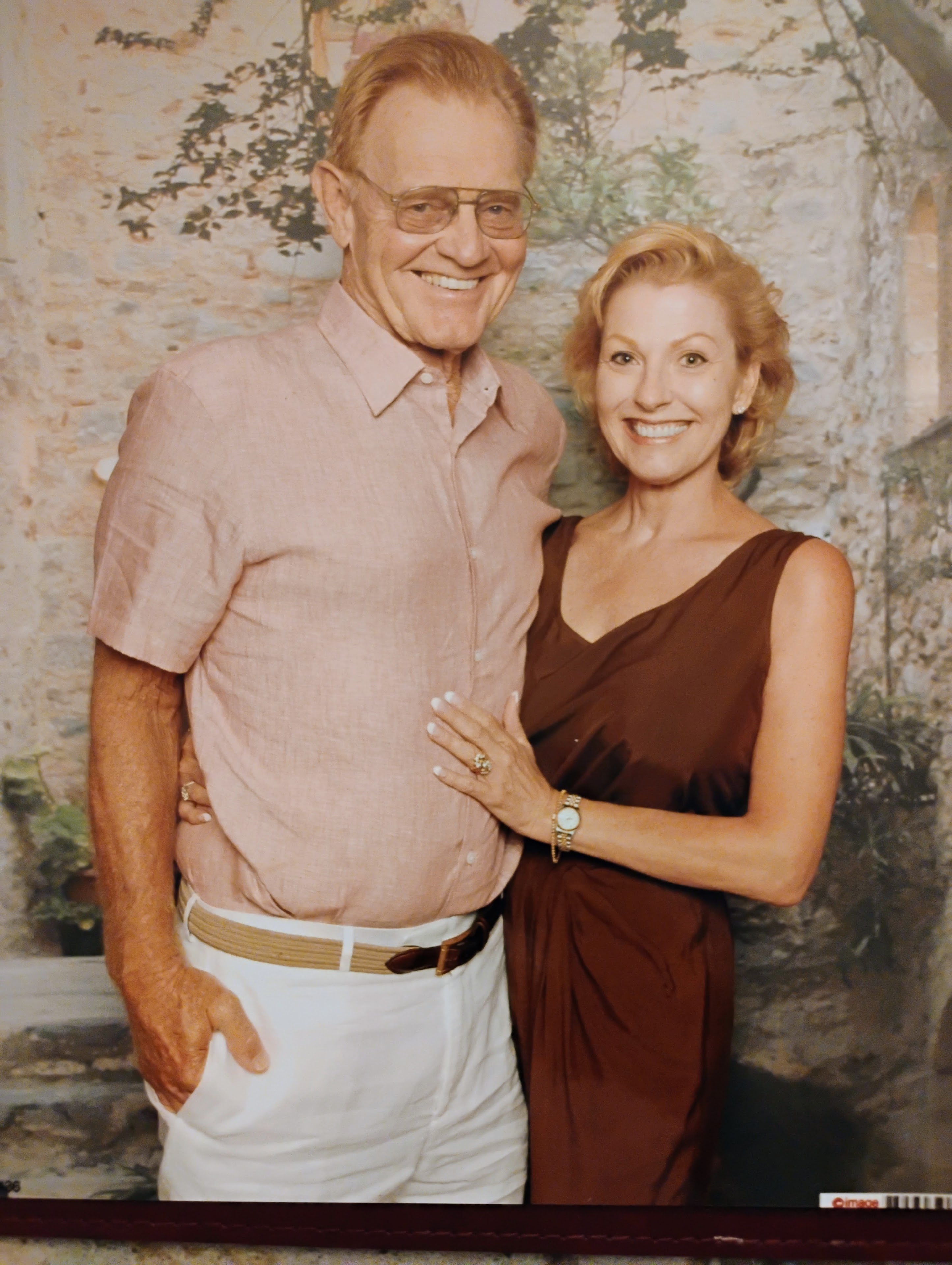 A man and woman stand close together, both smiling. They are in a garden with greenery around them.