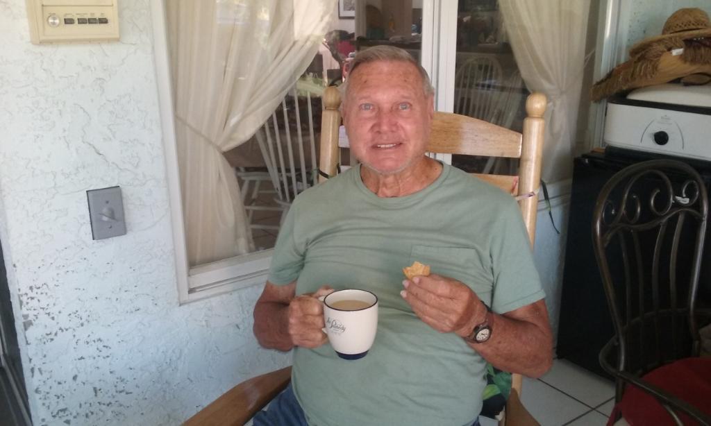 Man sits on a rocking chair holding a cup of coffee and a snack, relaxing on the porch.