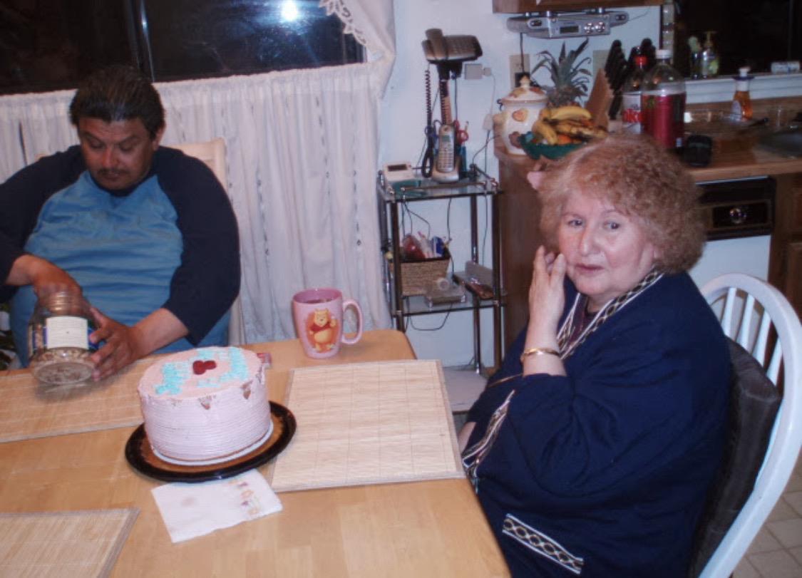 Two people are gathered at a table in a kitchen, with a cake present and a mug nearby.