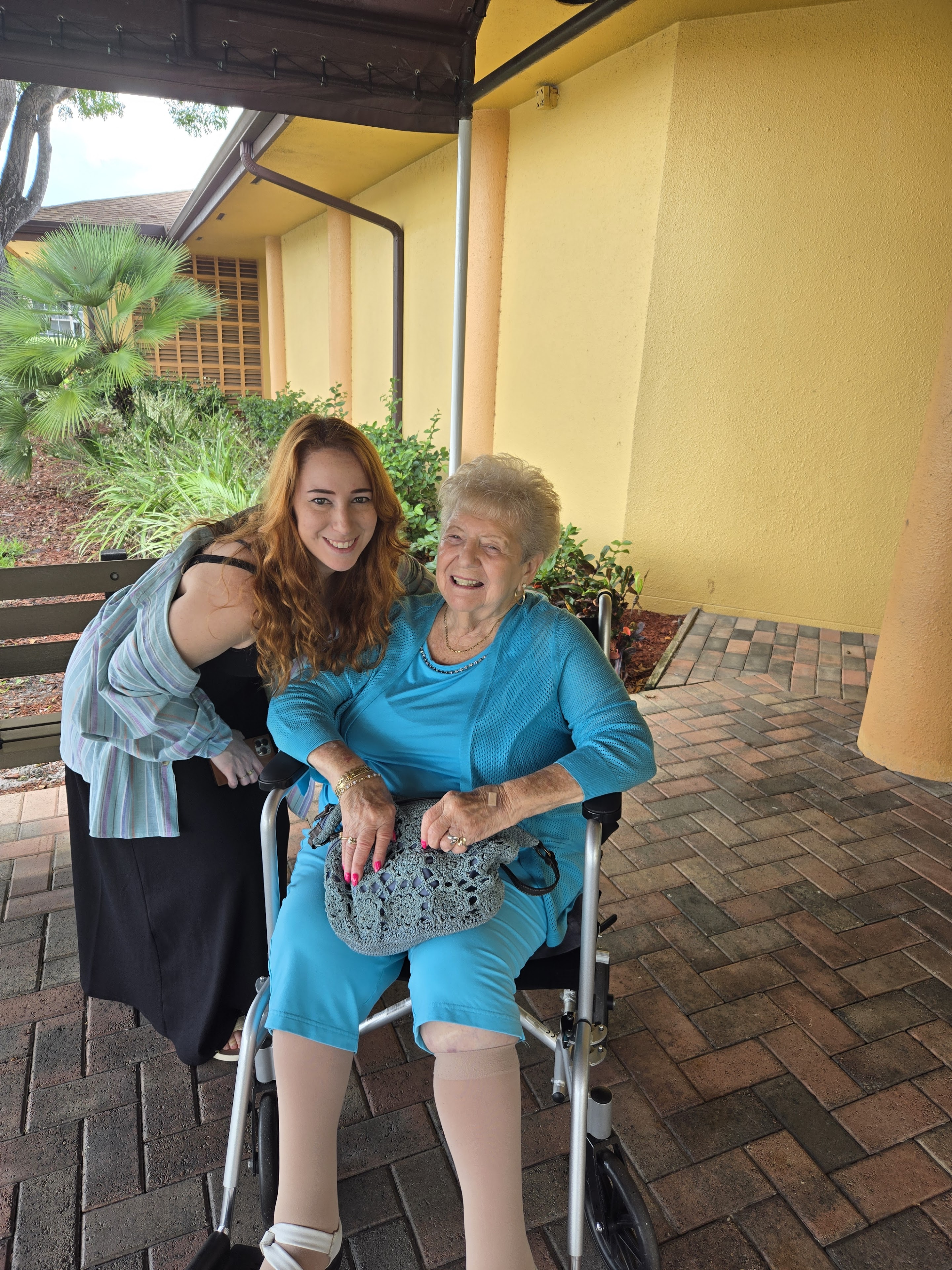 A young woman stands next to an elderly woman in a wheelchair. They both smile at the camera.