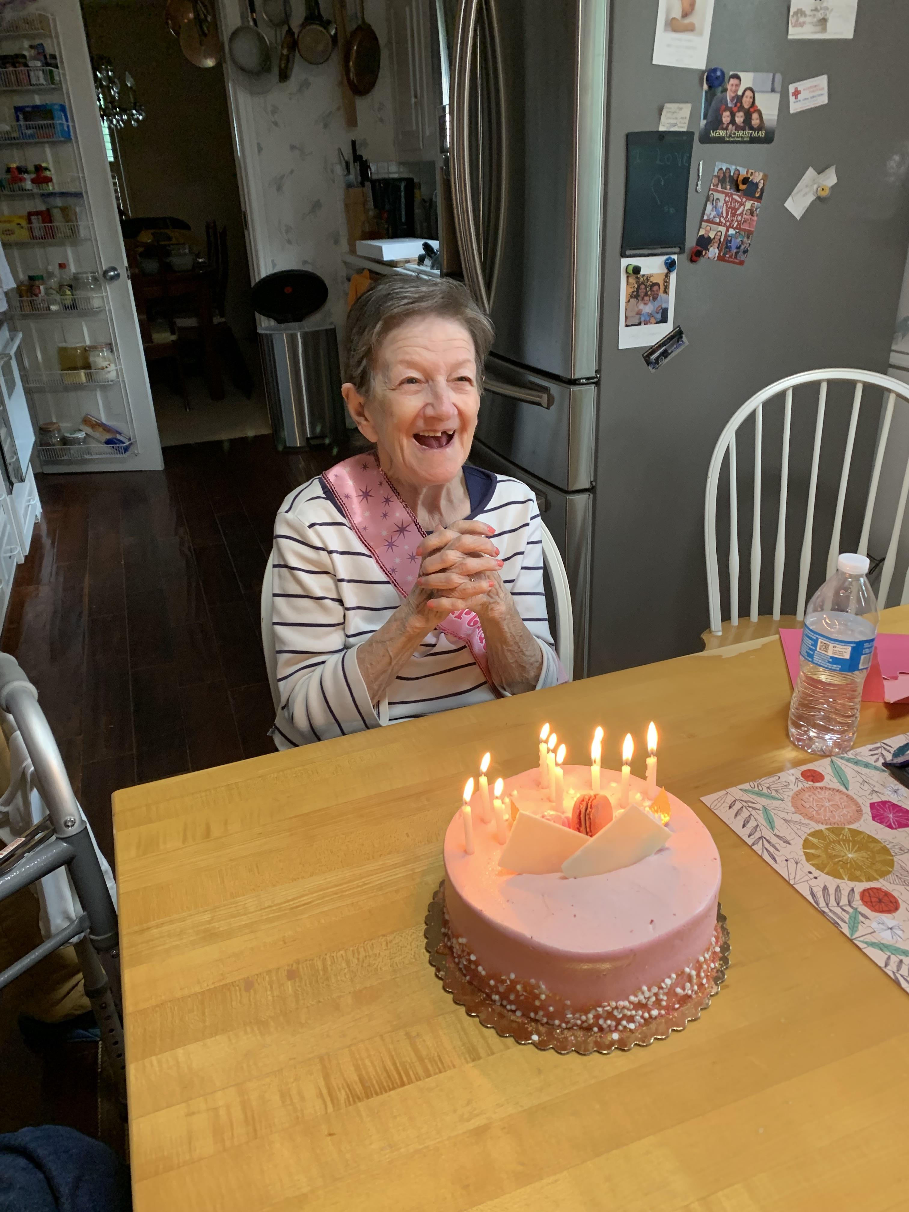 A woman sits at a table smiling while looking at a birthday cake with candles.