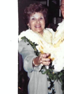Woman wearing flowers smiles widely as she gestures with her hand during a festive gathering.