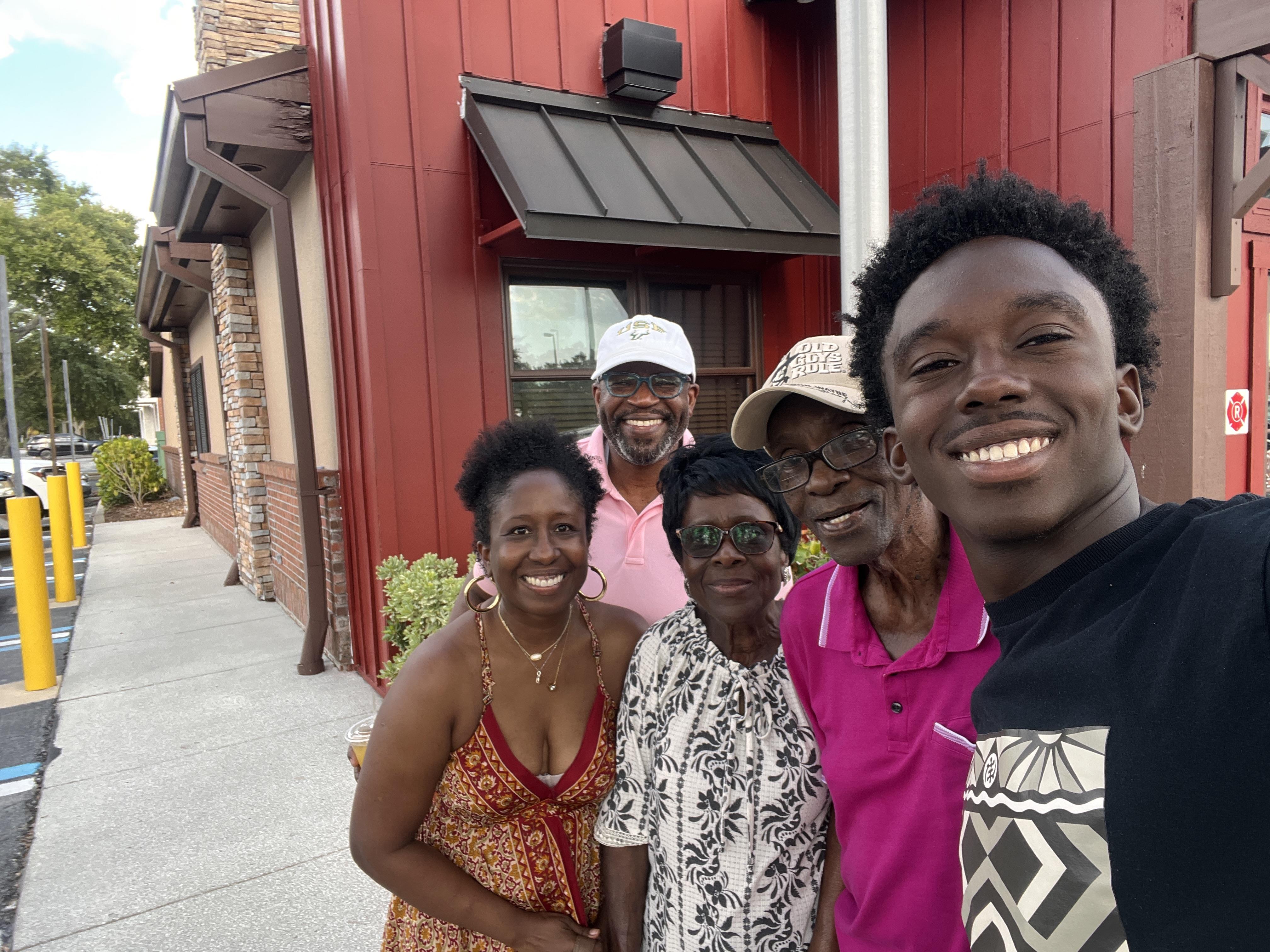 Five people stand close together and smile outside a red building on a sunny day.