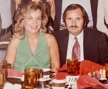 A couple sits together at a table with drinks, surrounded by others. They are at a social gathering.