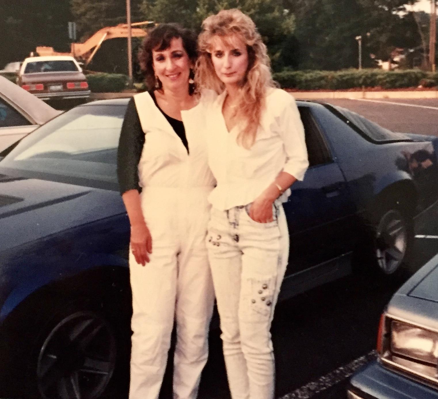 Two women pose in a parking lot by blue cars with a sunny backdrop in the daytime.