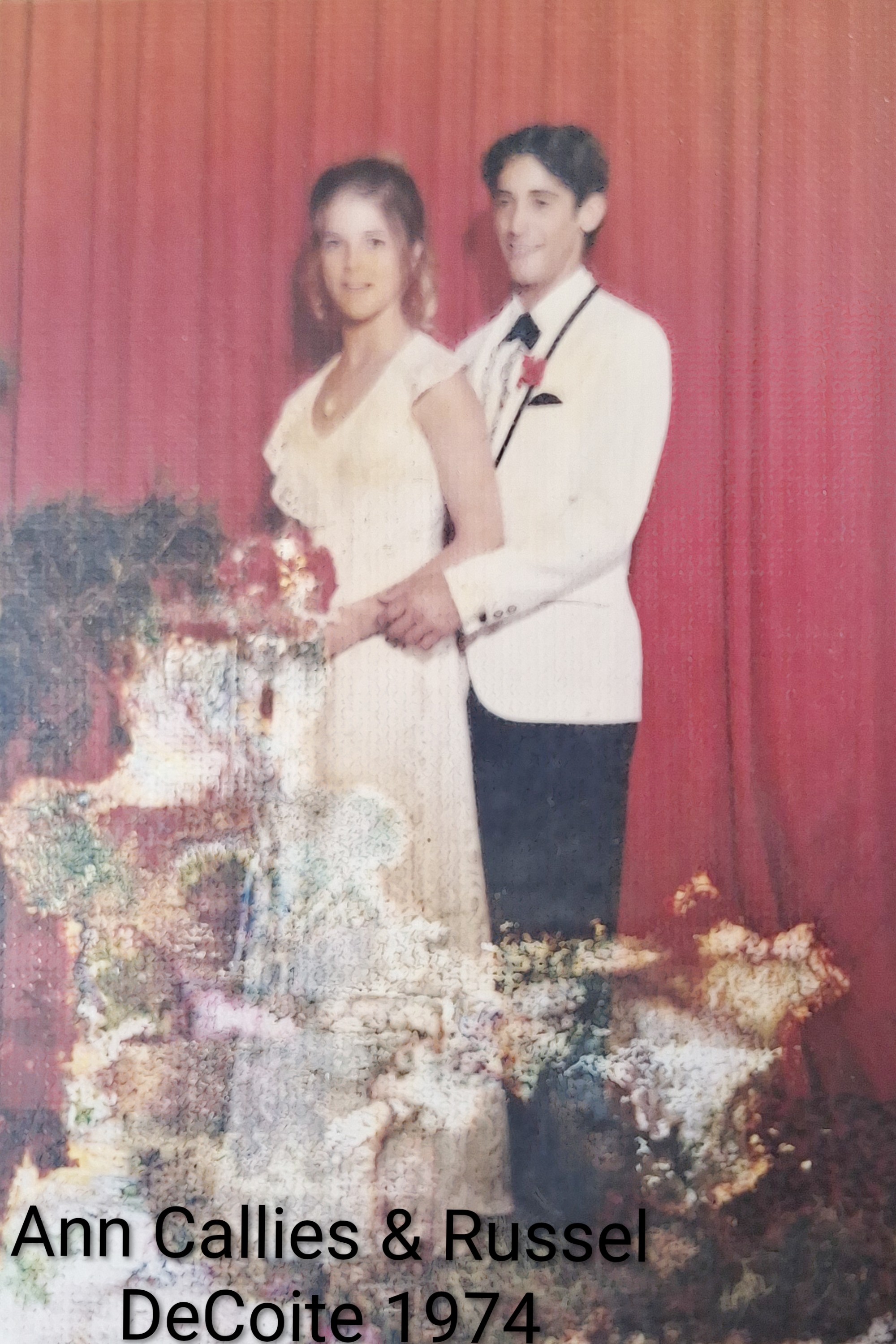 A couple in formal wear holds flowers, posing together in a studio with red curtains.