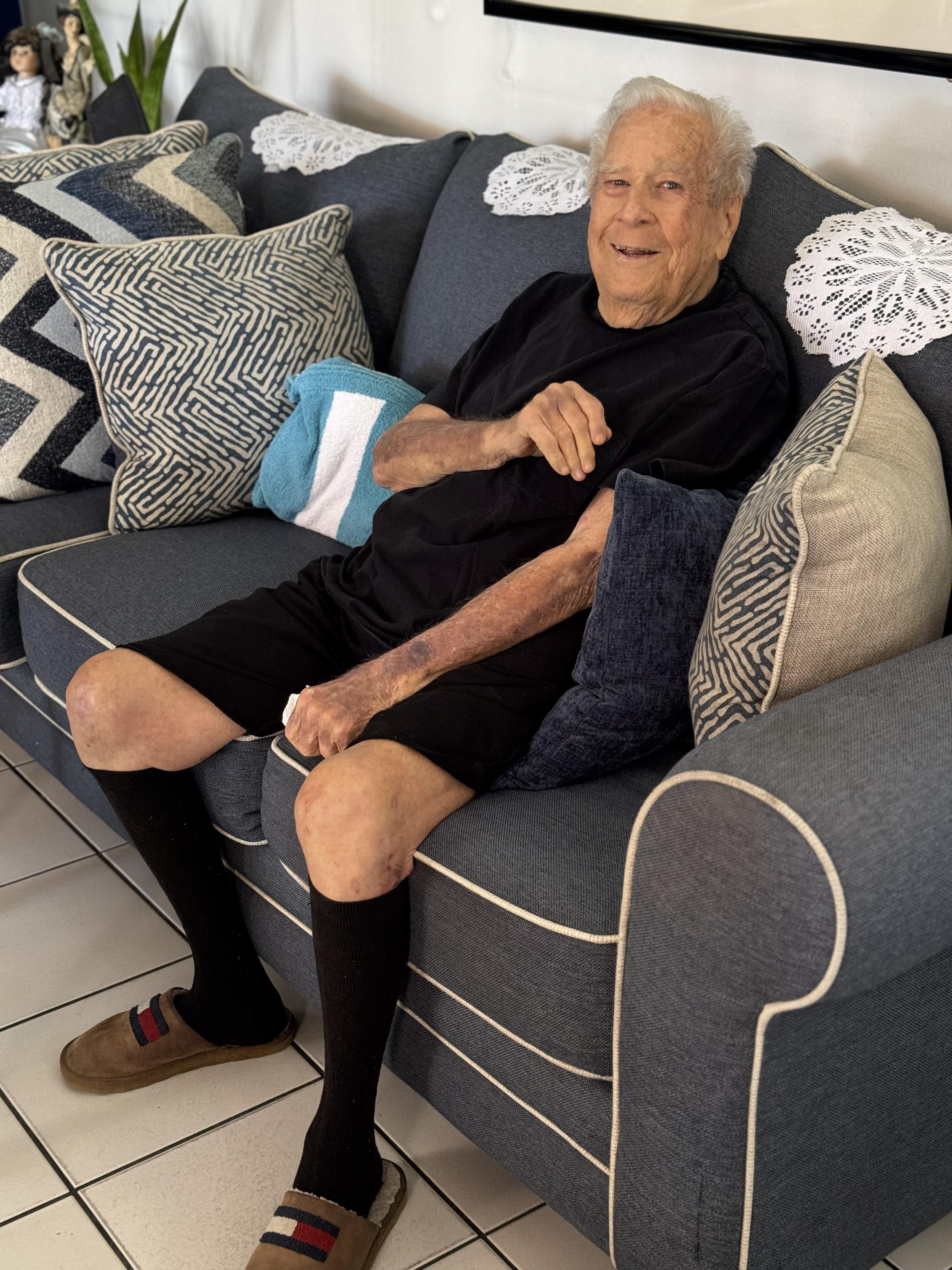 An elderly man is sitting comfortably on a couch with cushions.