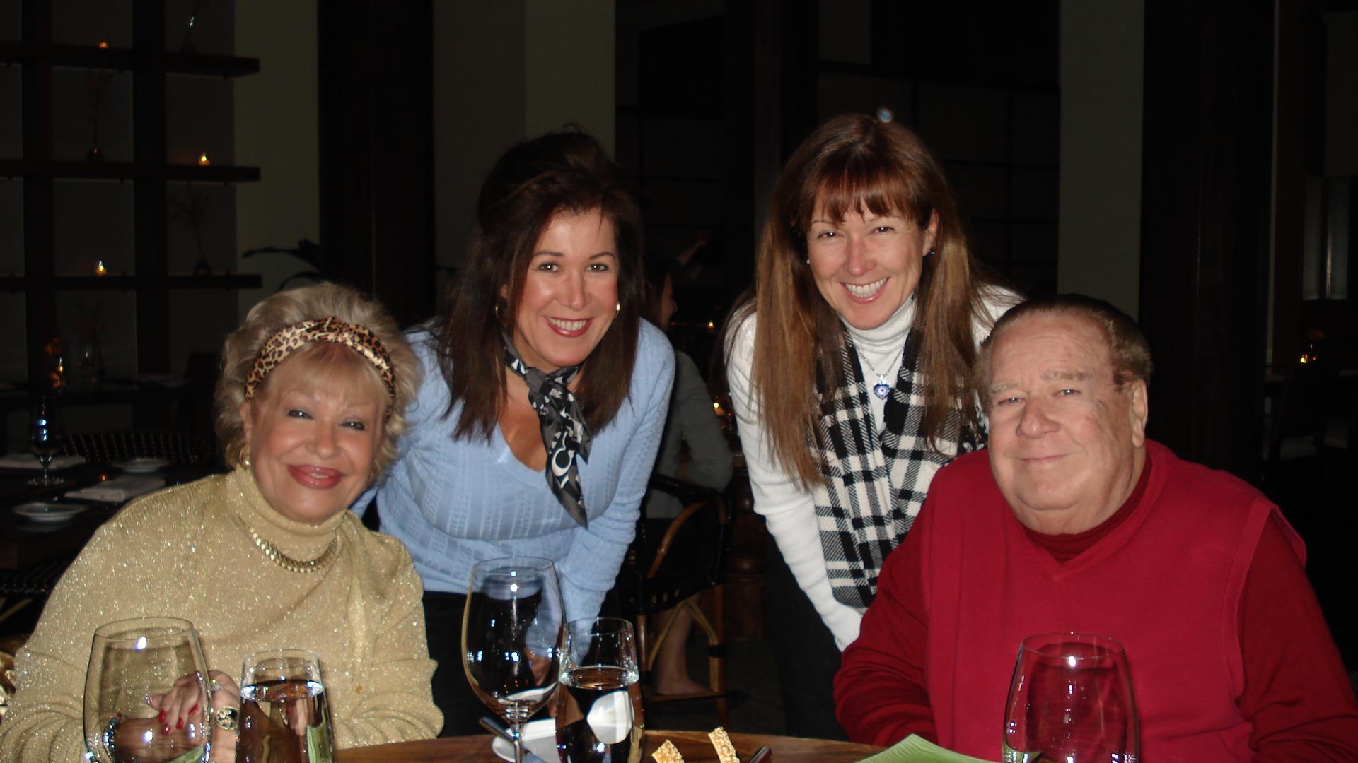 Four people smile at a table with drinks, enjoying a dinner gathering at night.