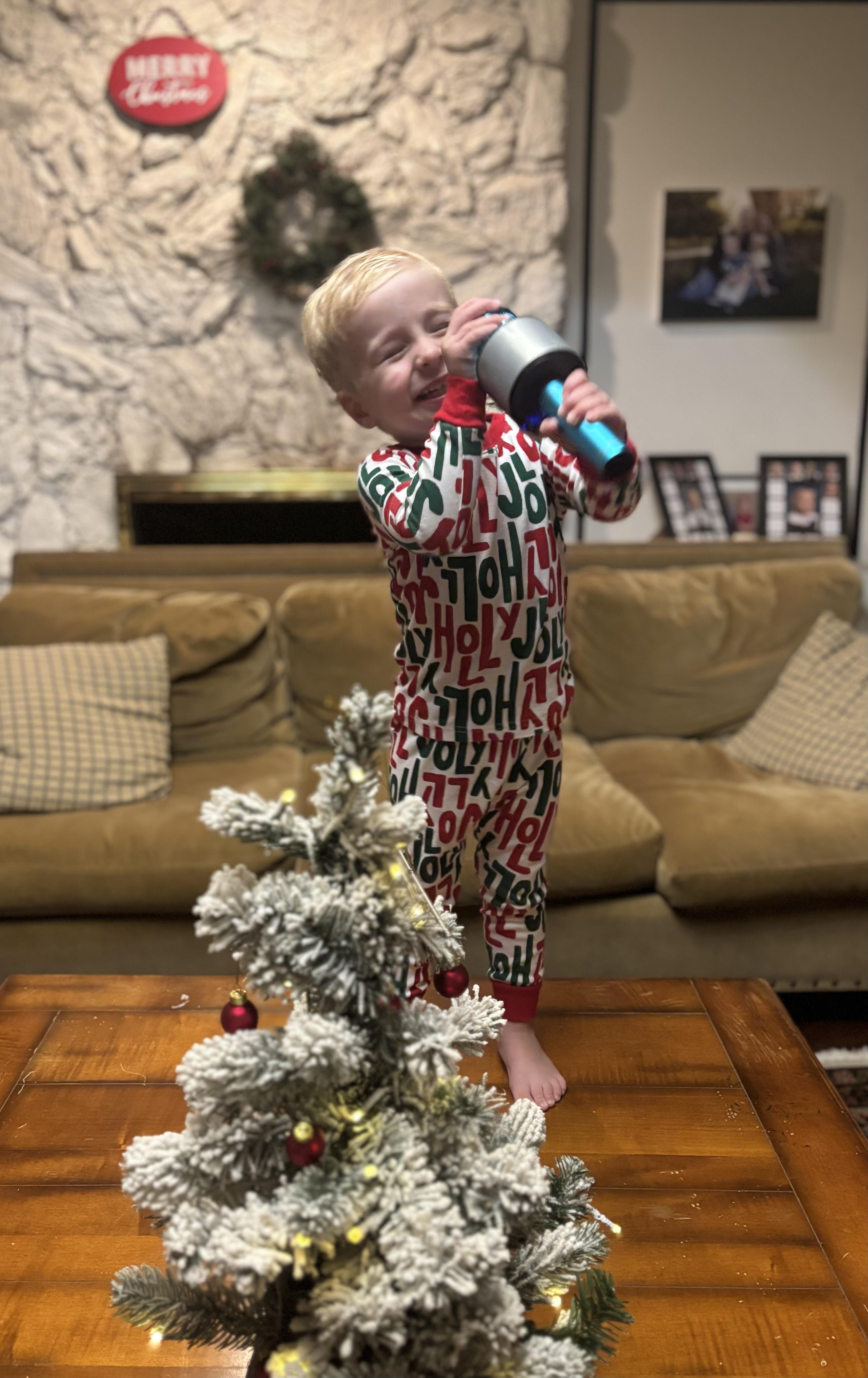 A child in festive pajamas sings with a microphone by a small Christmas tree.