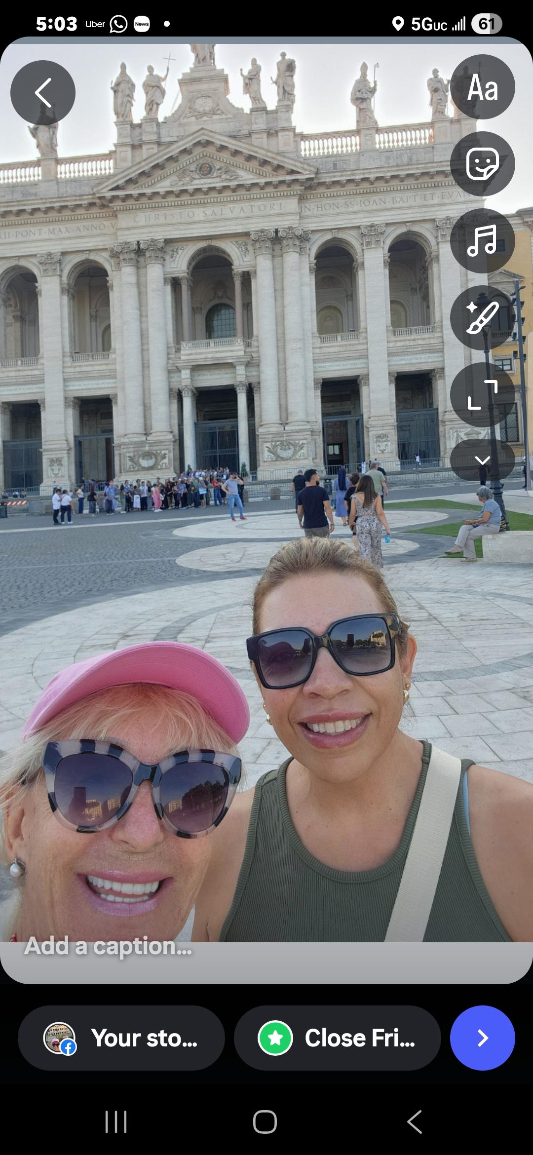 Two women smile together in front of a grand building with many people in the background.