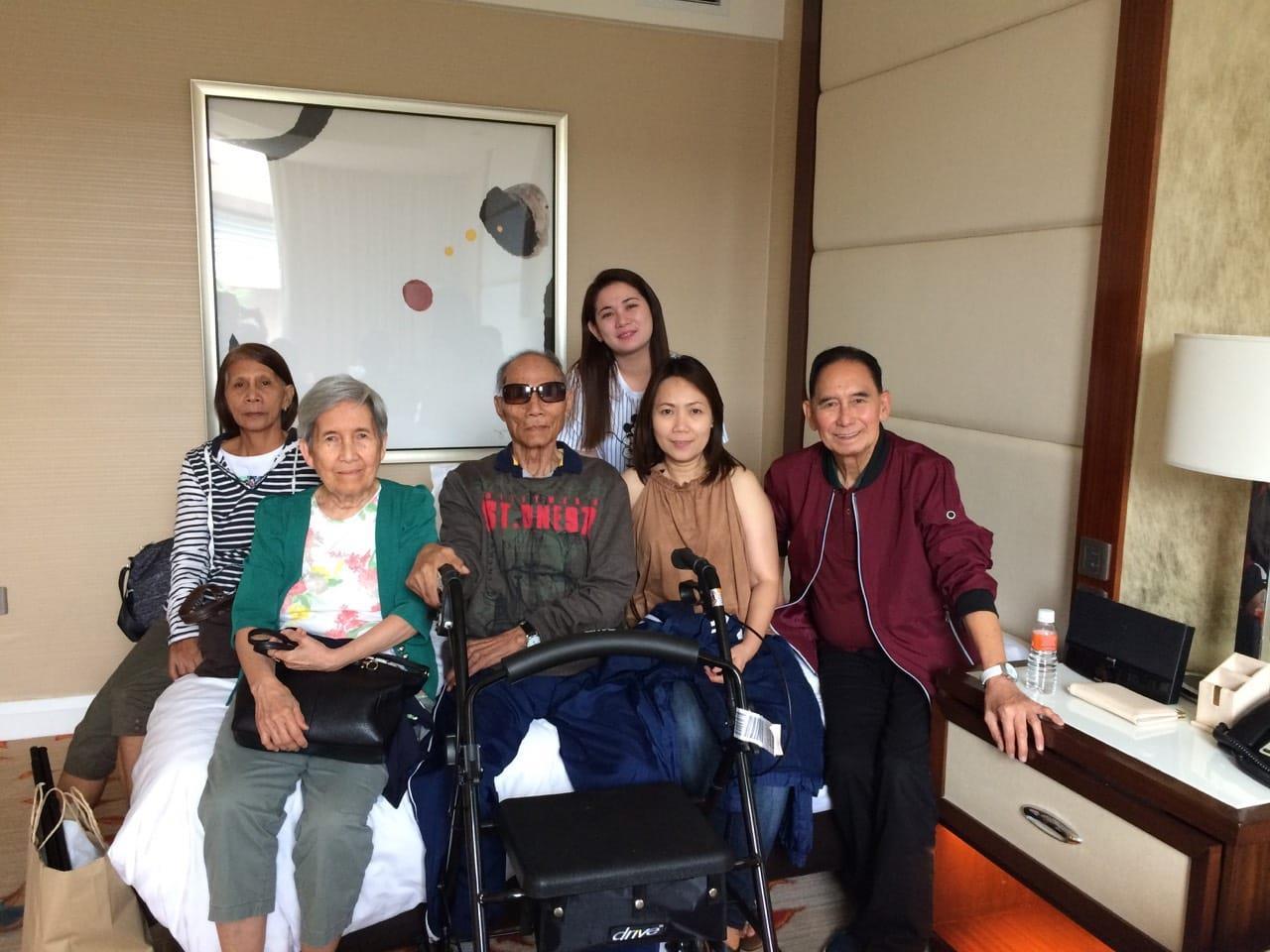 Family members spend time together in a hotel room with their elder relative and share smiles.