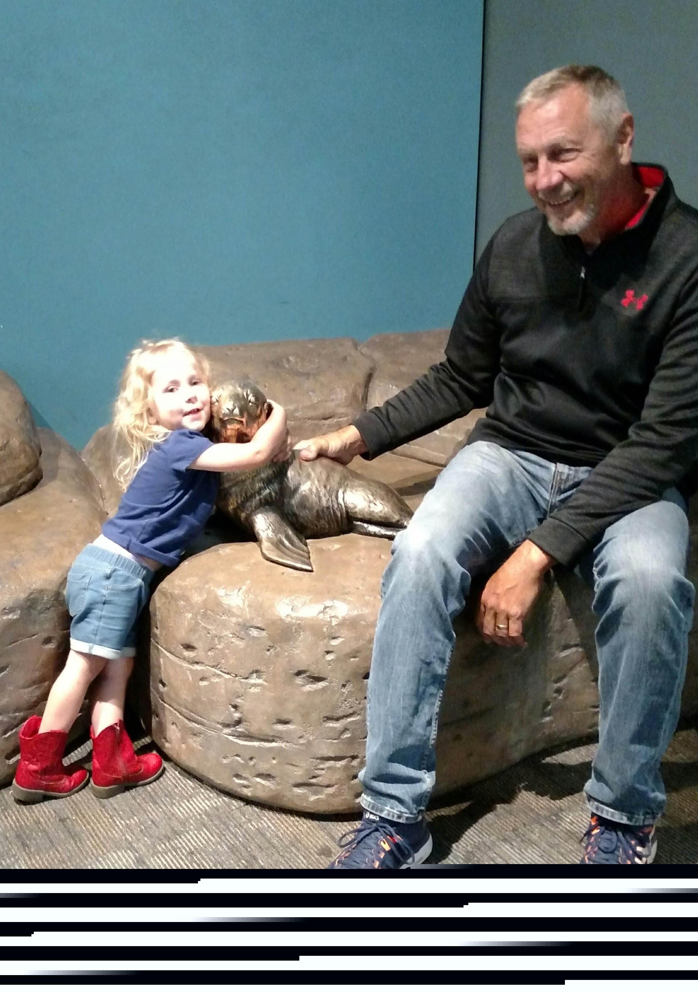 A young girl hugs a seal statue while a man smiles and sits on the couch beside her.