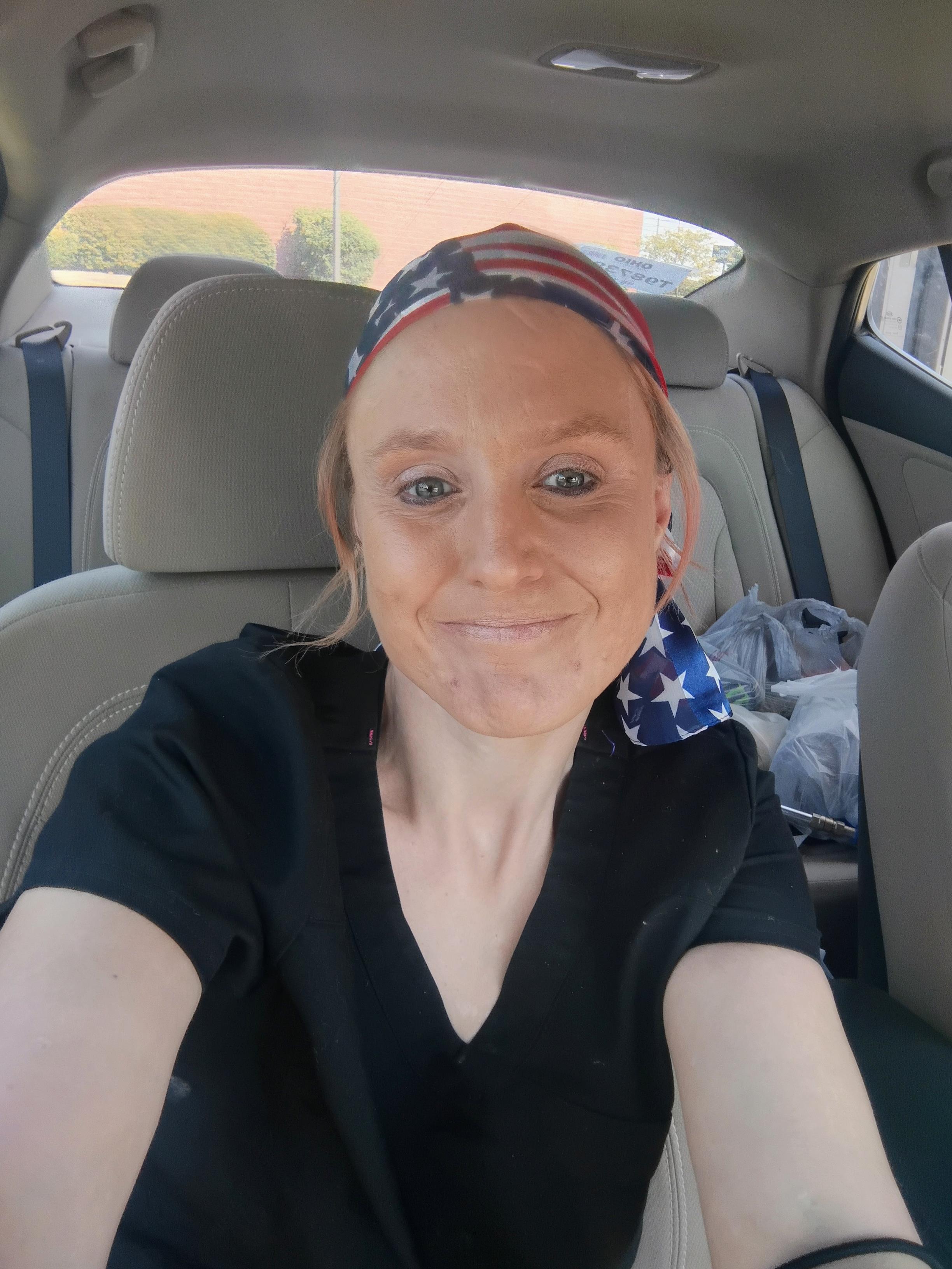 Woman sits in a vehicle, wearing a headscarf and smiling at the camera while parked.