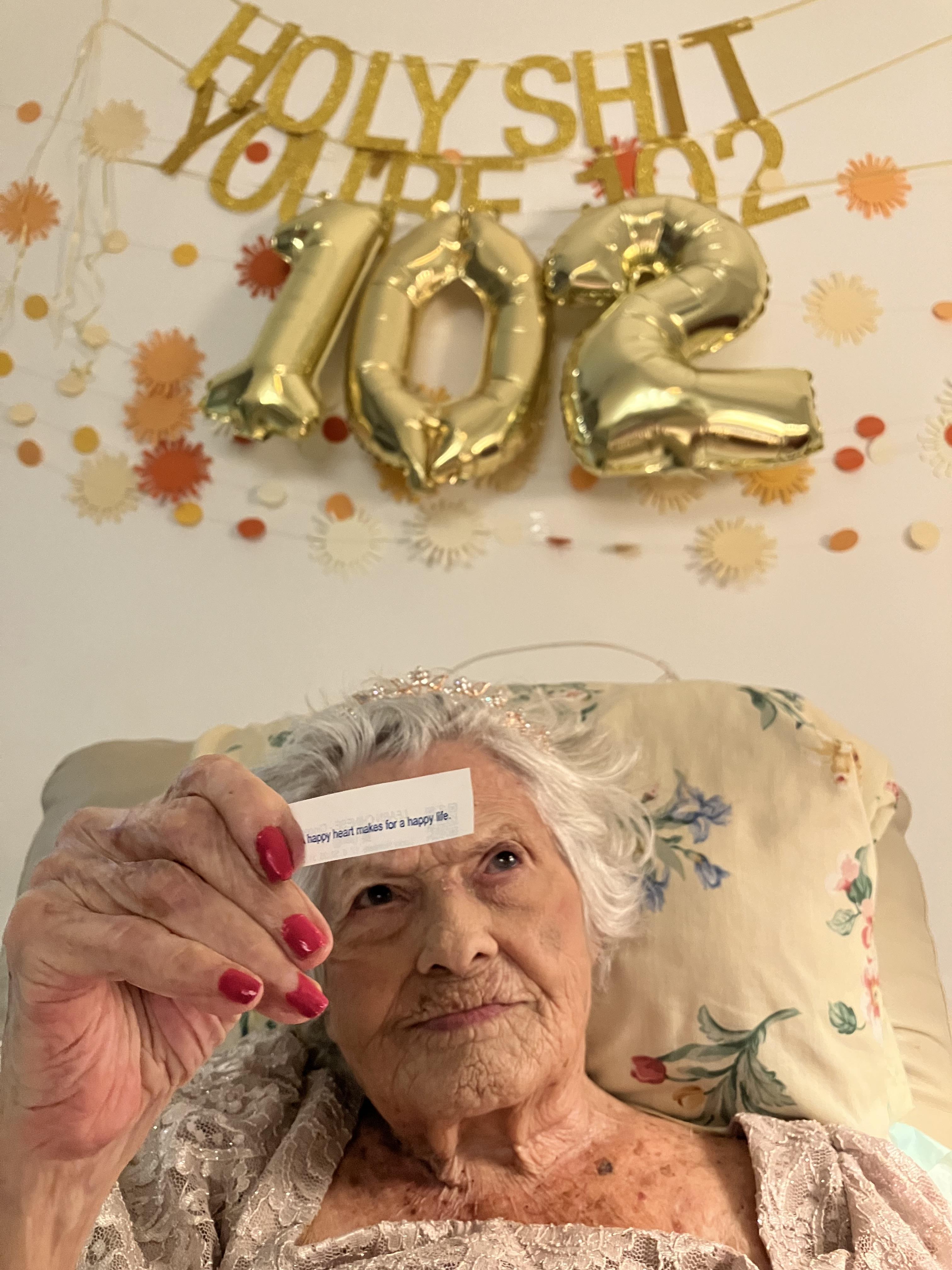 A woman holds a card while celebrating her 102nd birthday with friends and decorations.