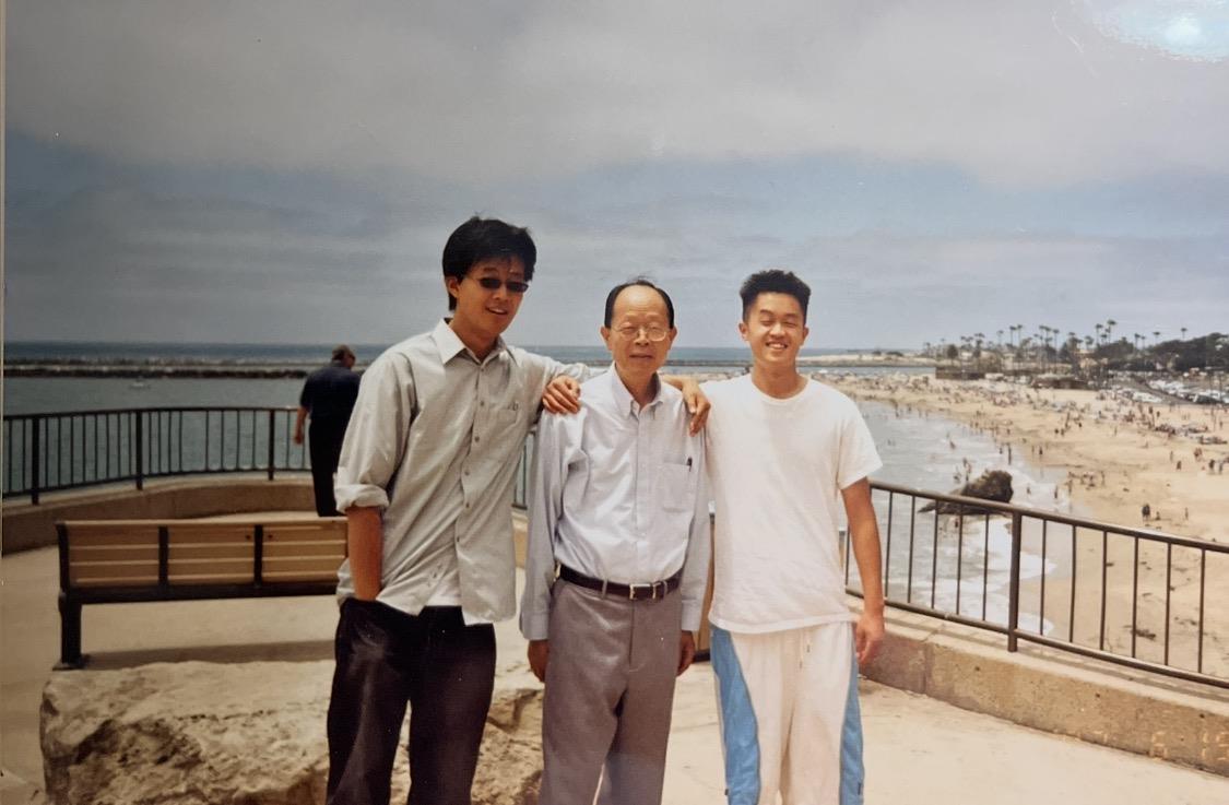 Three people pose outside near the ocean with a clear sky above and a crowd in the background.