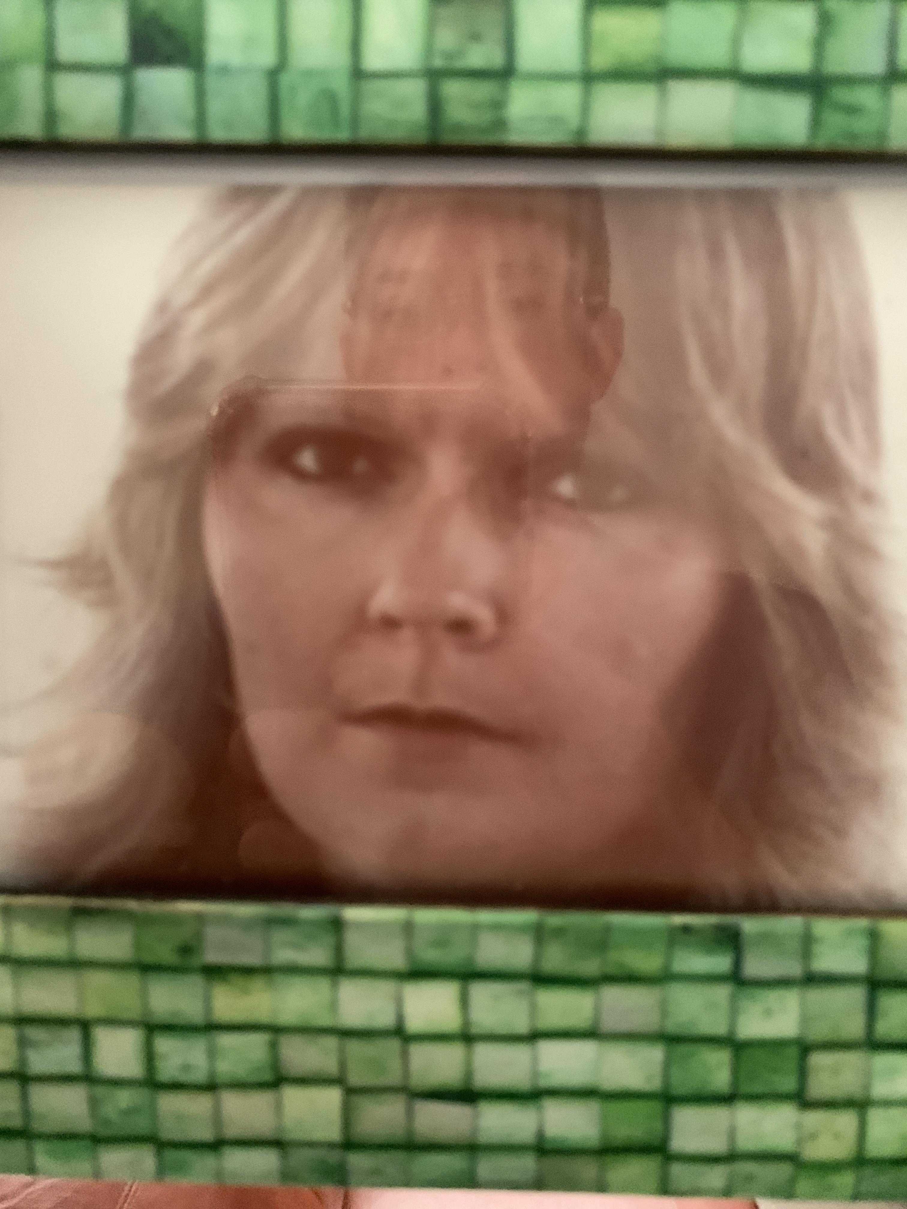 A woman with long hair stares at the camera. She stands in front of a green tiled surface.