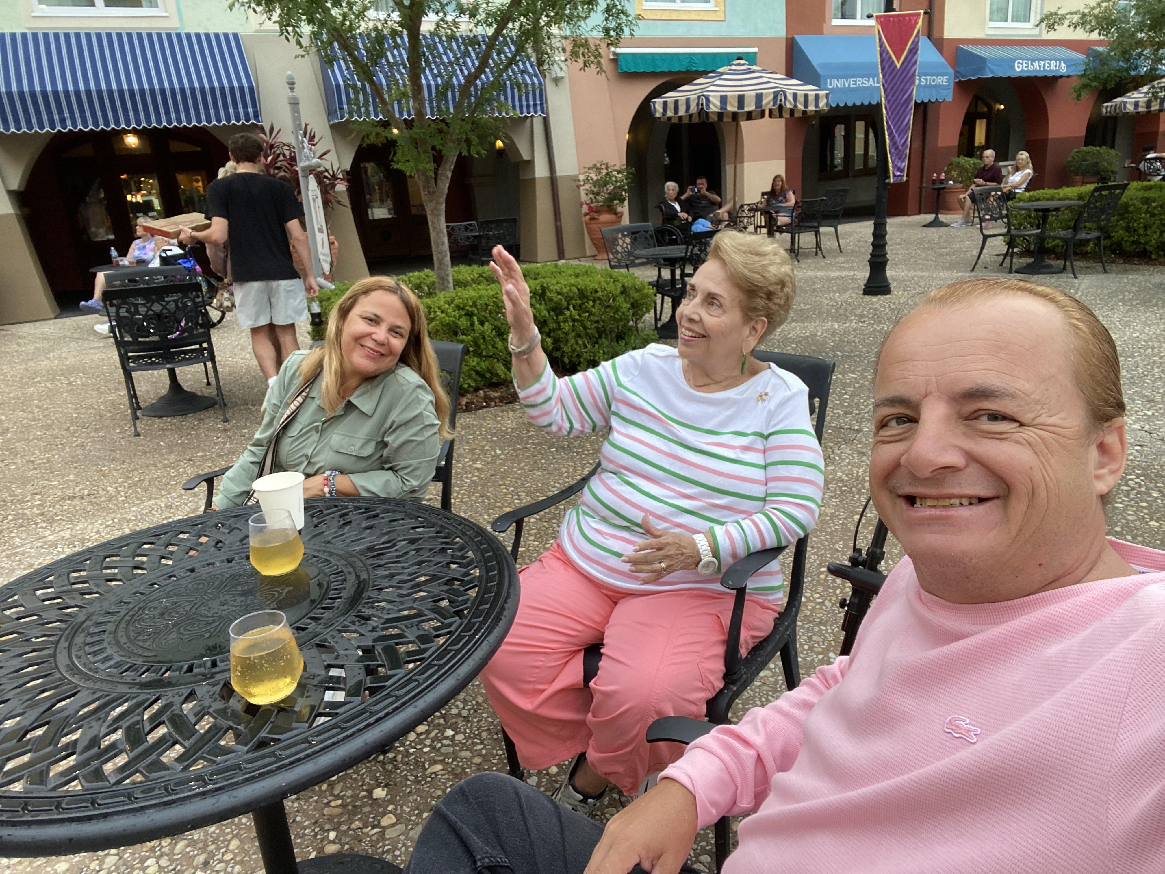 Three people share a fun time at a plaza with drinks placed on the table in the afternoon sun.