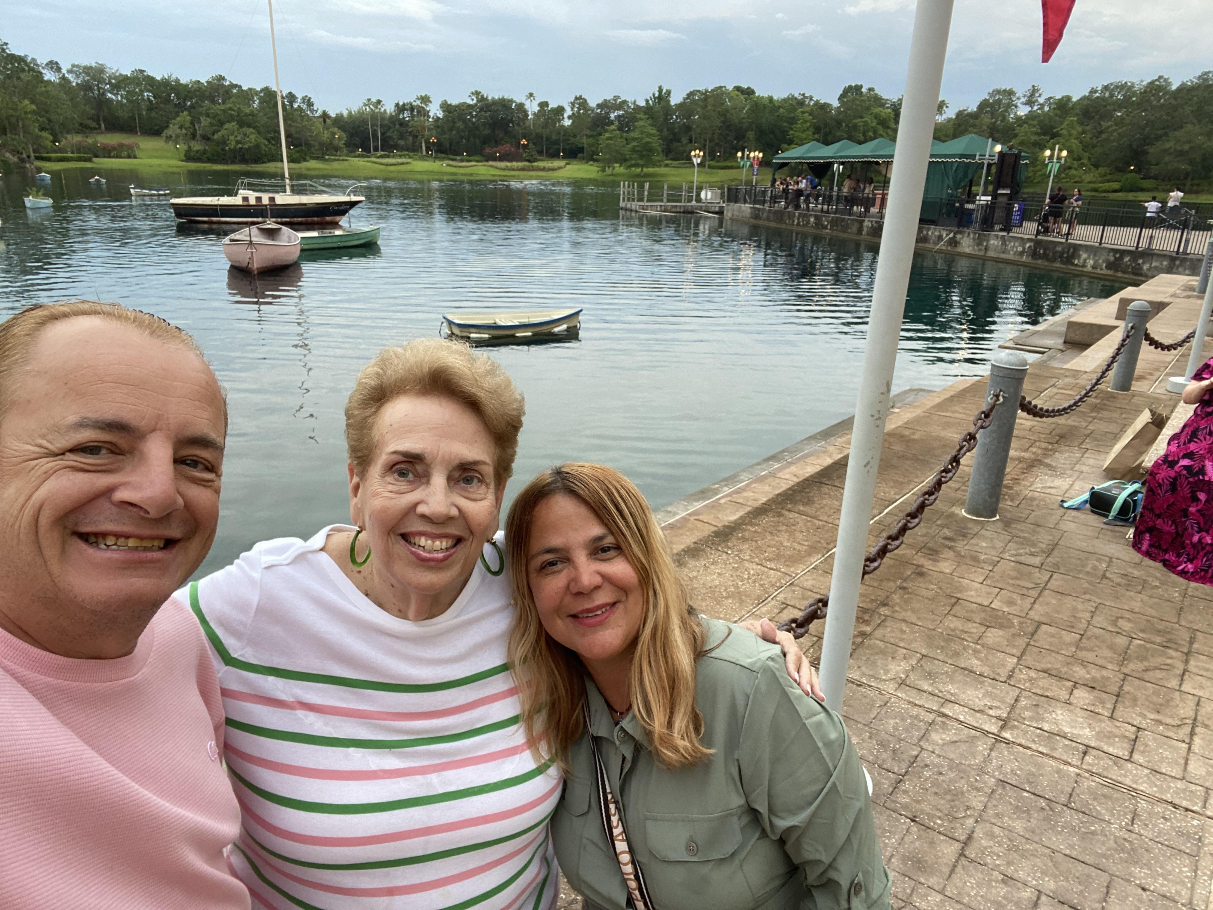 Three women smile together by a lake with boats floating nearby and trees in the distance.