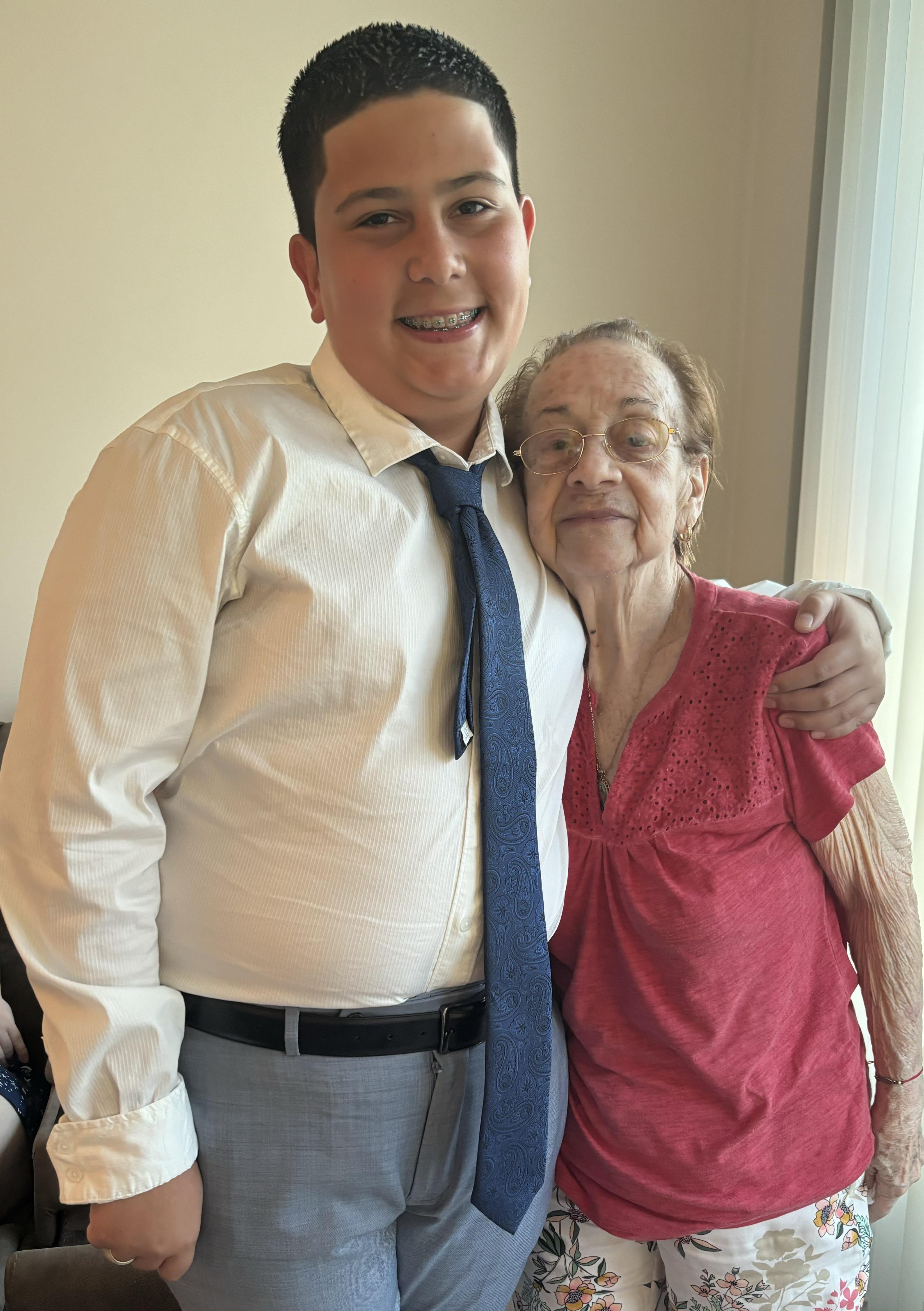 A young man stands next to an elderly woman with a smile, both enjoying a joyful moment indoors.