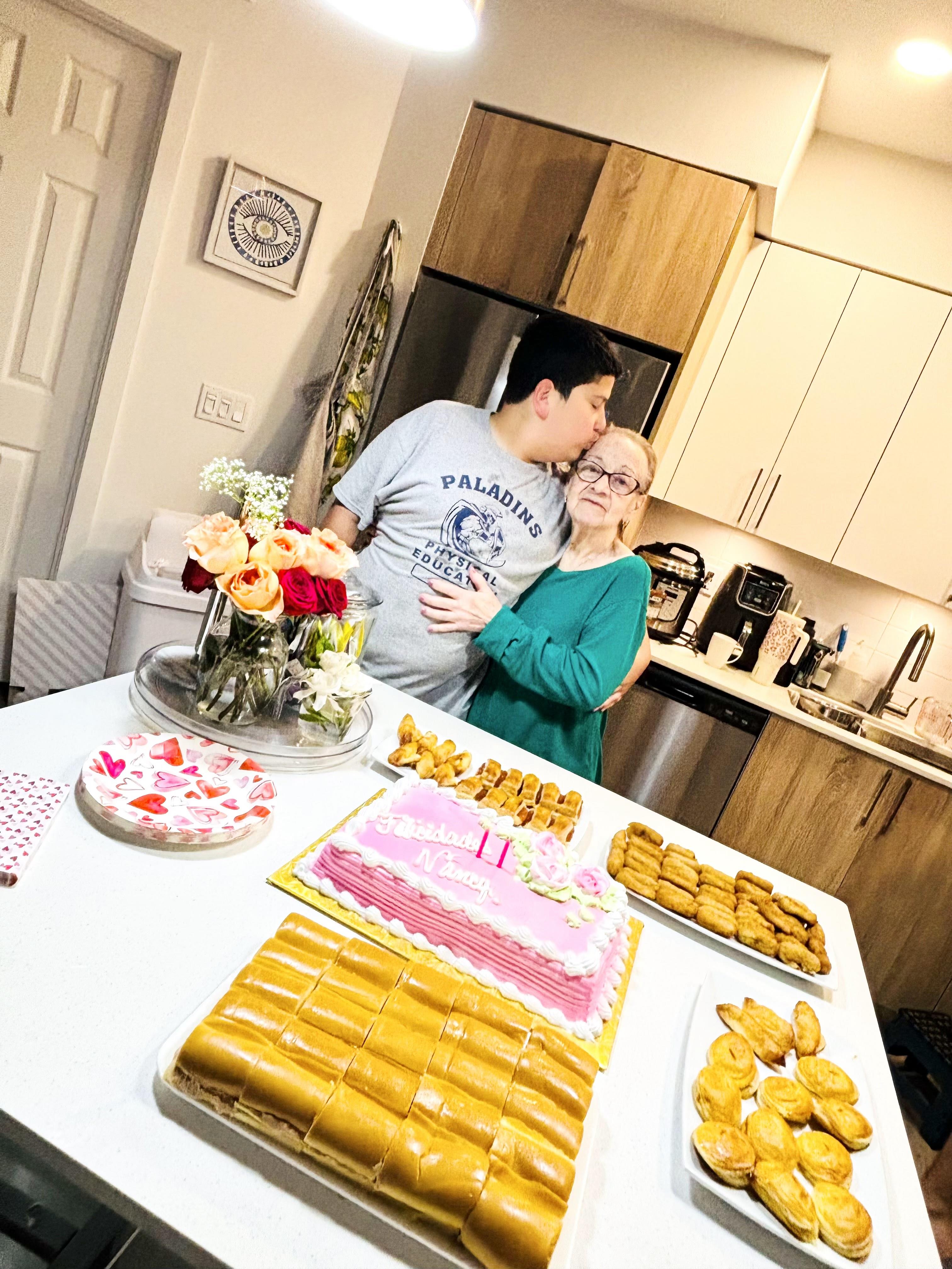A couple stands close together in a kitchen filled with snacks and a large cake on the table.